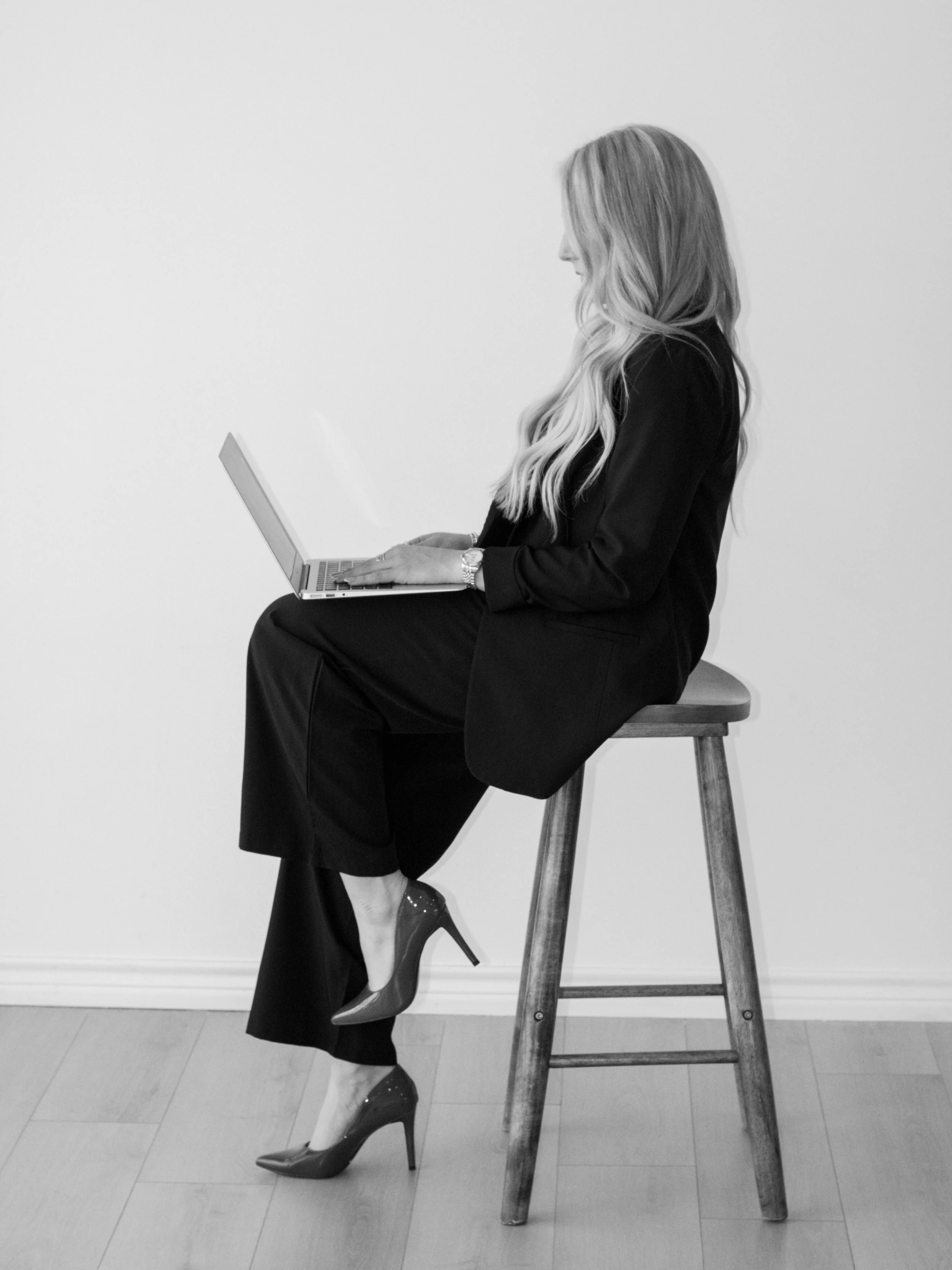 A woman with long wavy hair sitting sideways on a wooden stool, wearing high heels and a black outfit, working on a laptop.