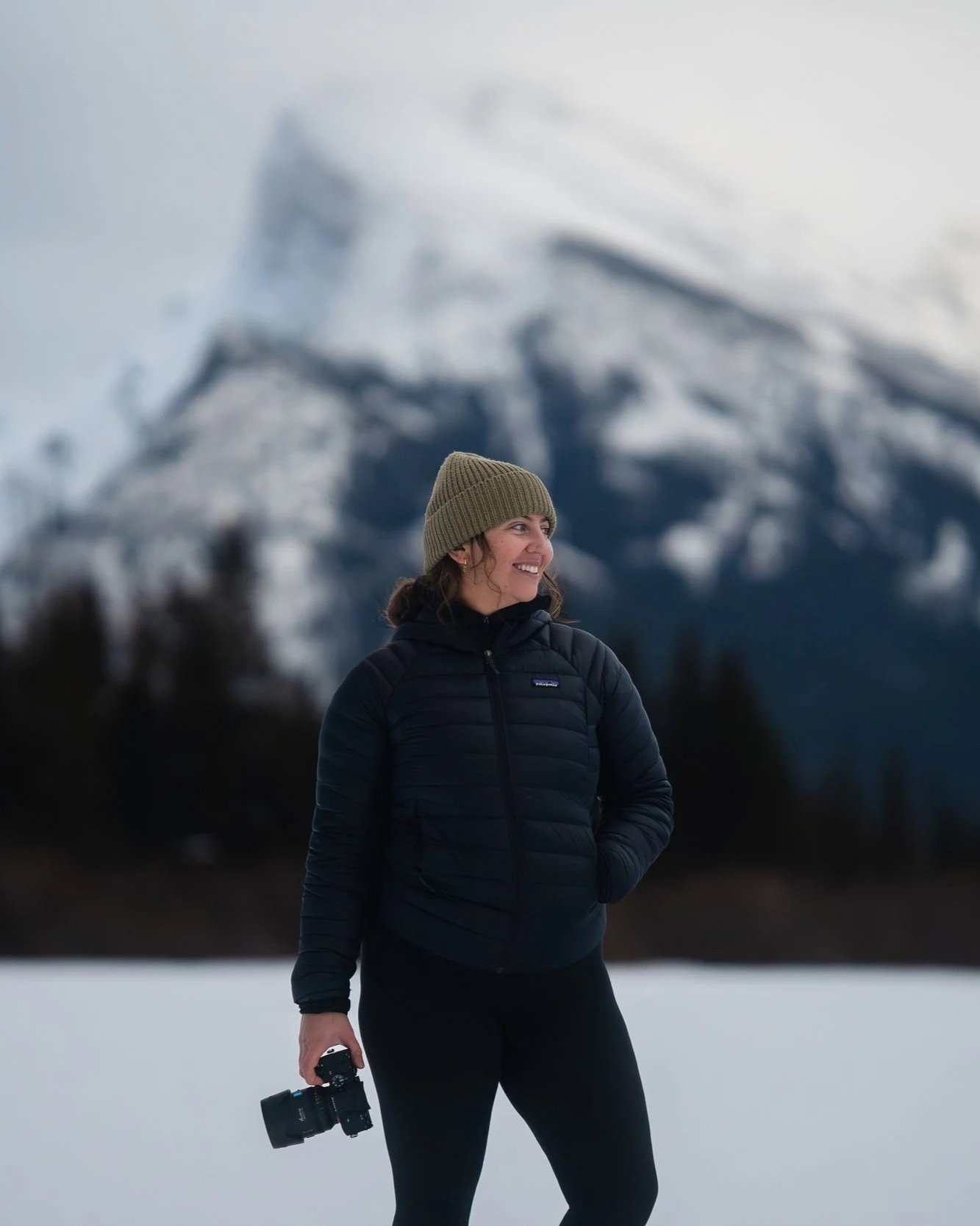 Female Canadian Photographer standing outdoors in a snowy landscape with snow-covered rocky mountains in the background, holding a camera, wearing a knit hat and a black puffy jacket, smiling.