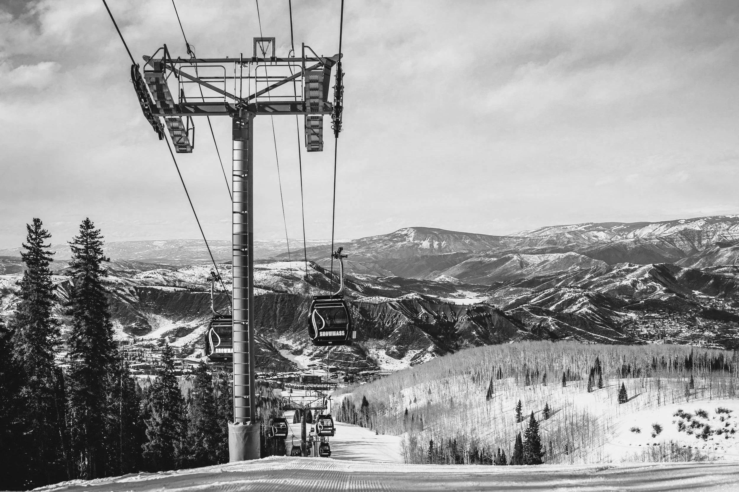 Black and white photo of a ski lift with empty chairs, snow-covered landscape, pine trees, and mountain range in the background.