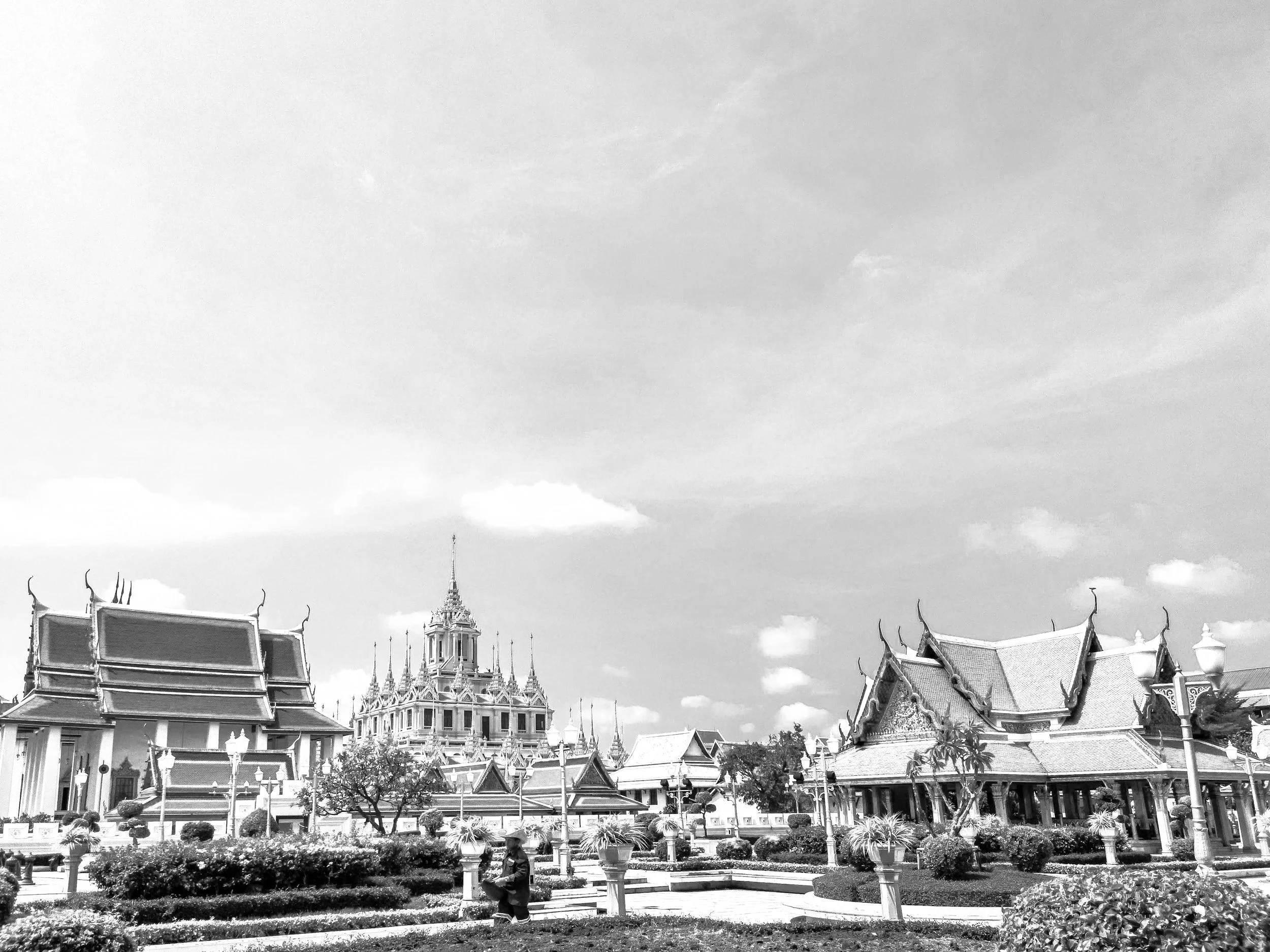 Traditional Thai temple complex with ornate buildings, a central tower, and landscaped garden, under a partly cloudy sky.