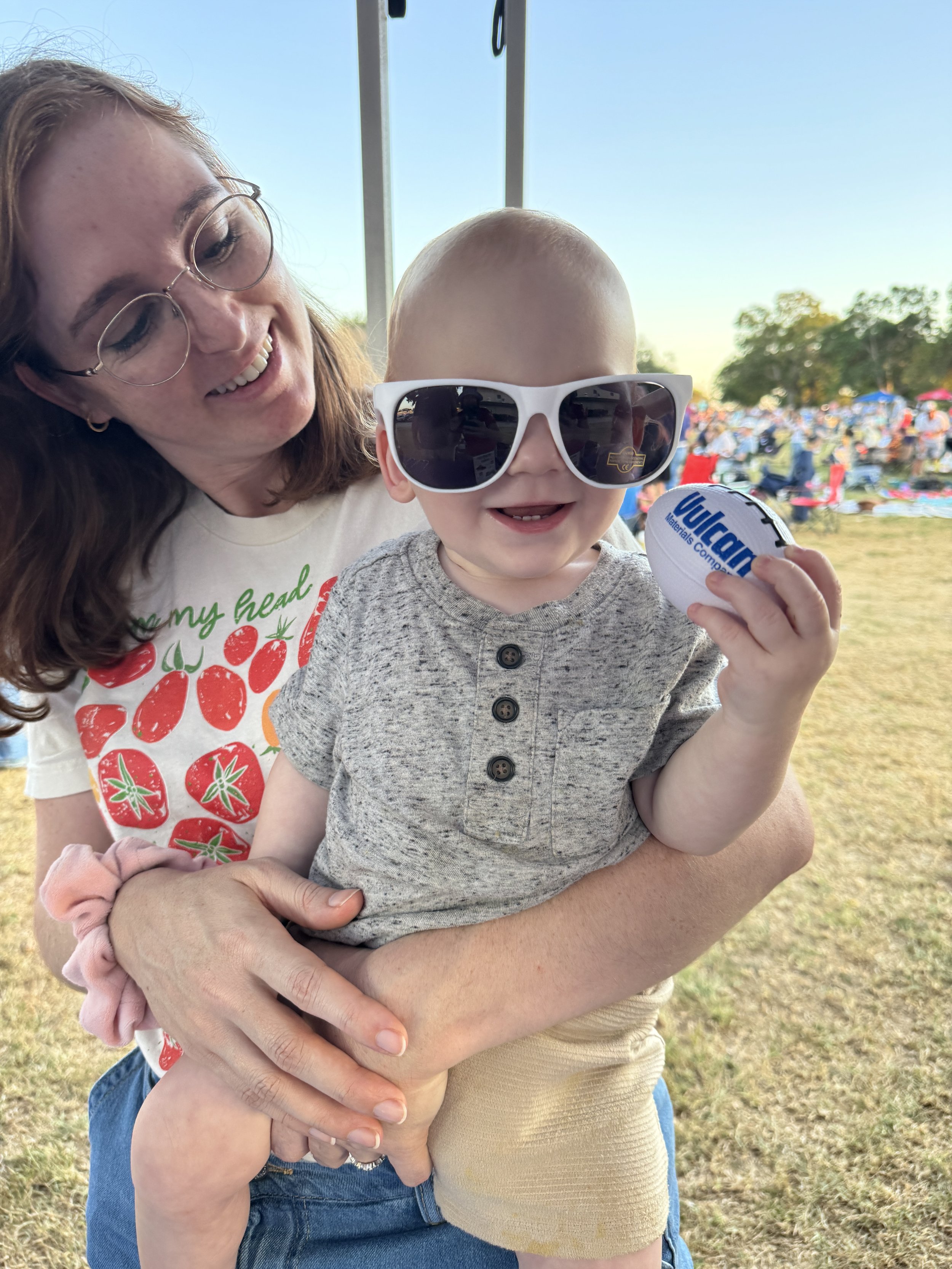 A woman holding a baby at an outdoor event with chairs and people in the background. The baby is wearing sunglasses and holding a football, smiling. The woman is smiling and wearing glasses.