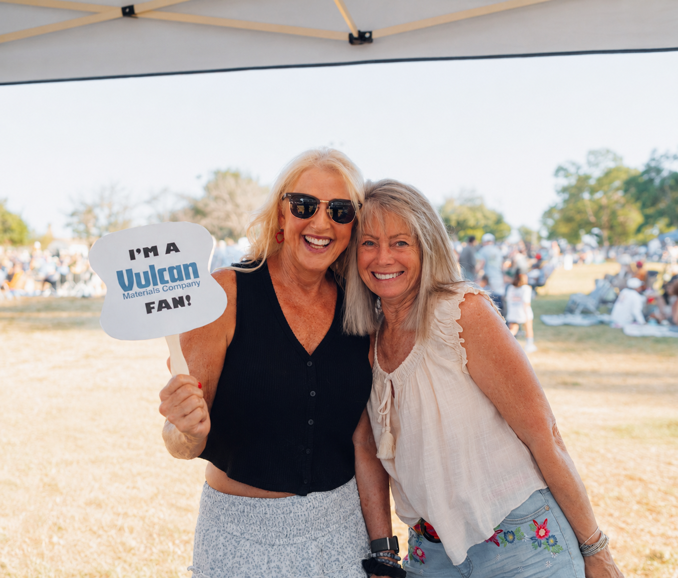 Two smiling women pose at an outdoor community event, with one holding a sign that reads “I’m a Vulcan Materials Company Fan!”