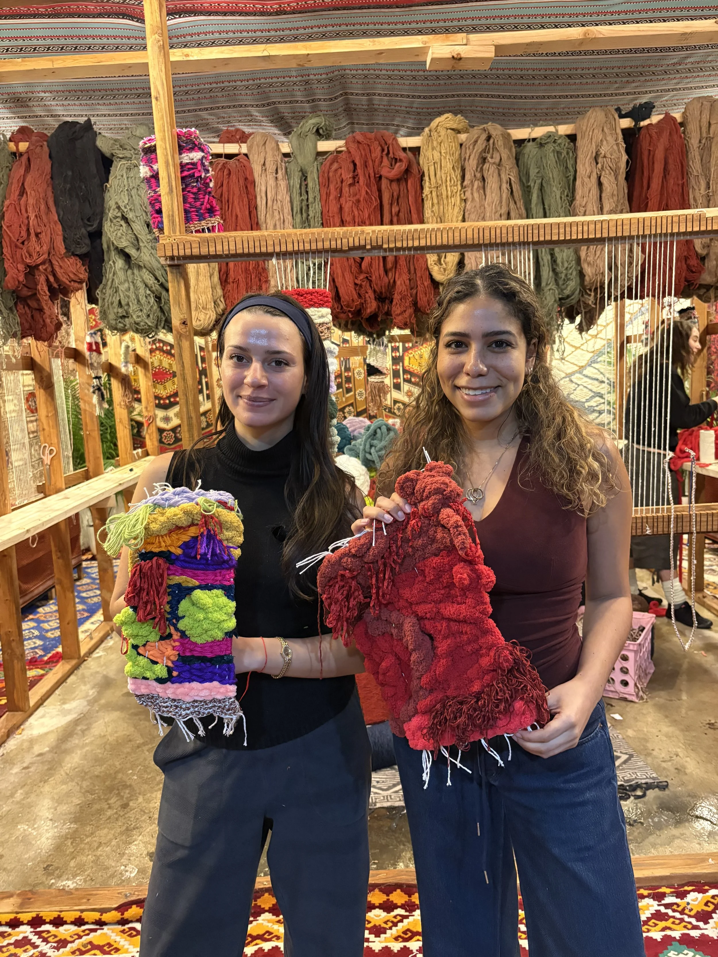 Two women standing in front of a display of colorful woven scarves and textiles, holding hand-knit or woven items in a market or craft fair setting.