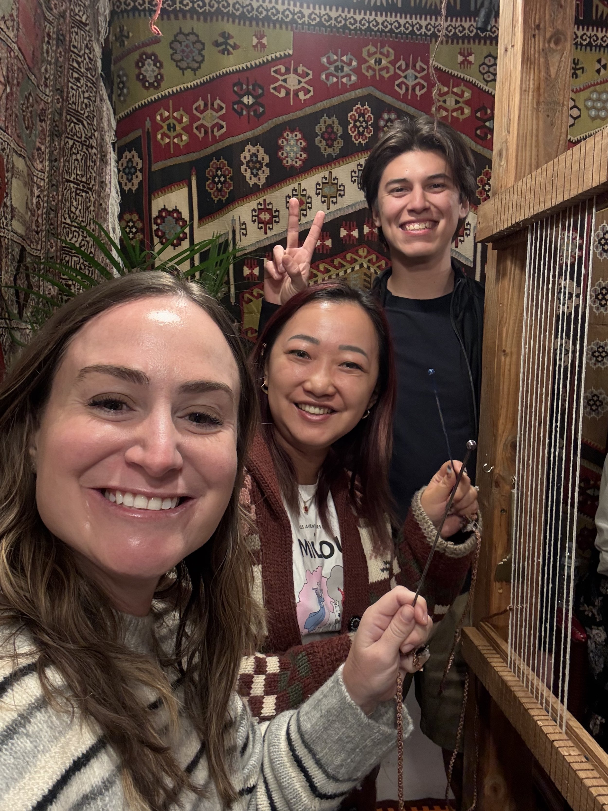 Three people smiling, taking a selfie in a room with colorful, patterned rugs hanging on the walls.