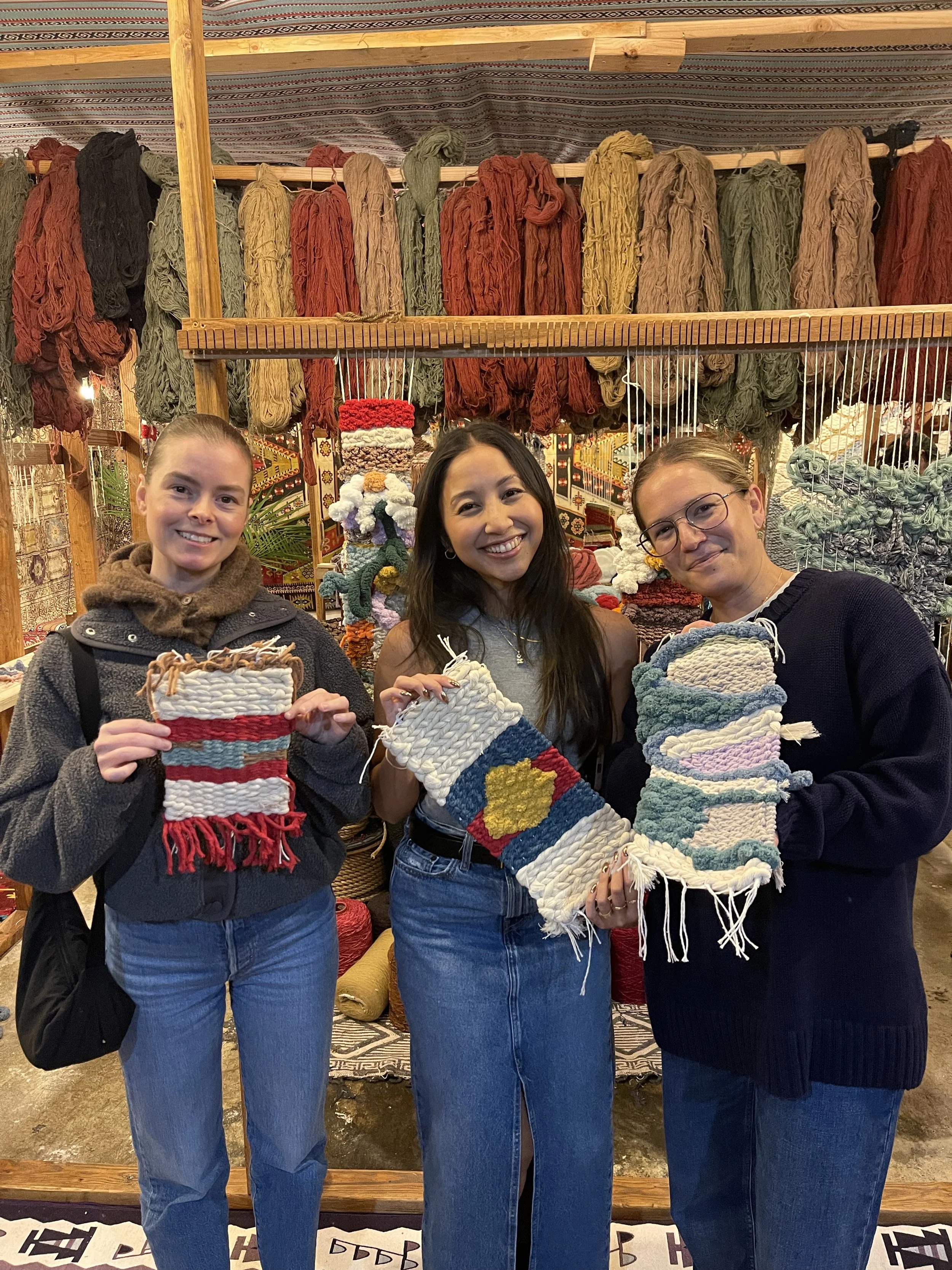 Three women are holding colorful woven slippers in a craft market. They are smiling and standing in front of shelves filled with yarn of various colors and textures.