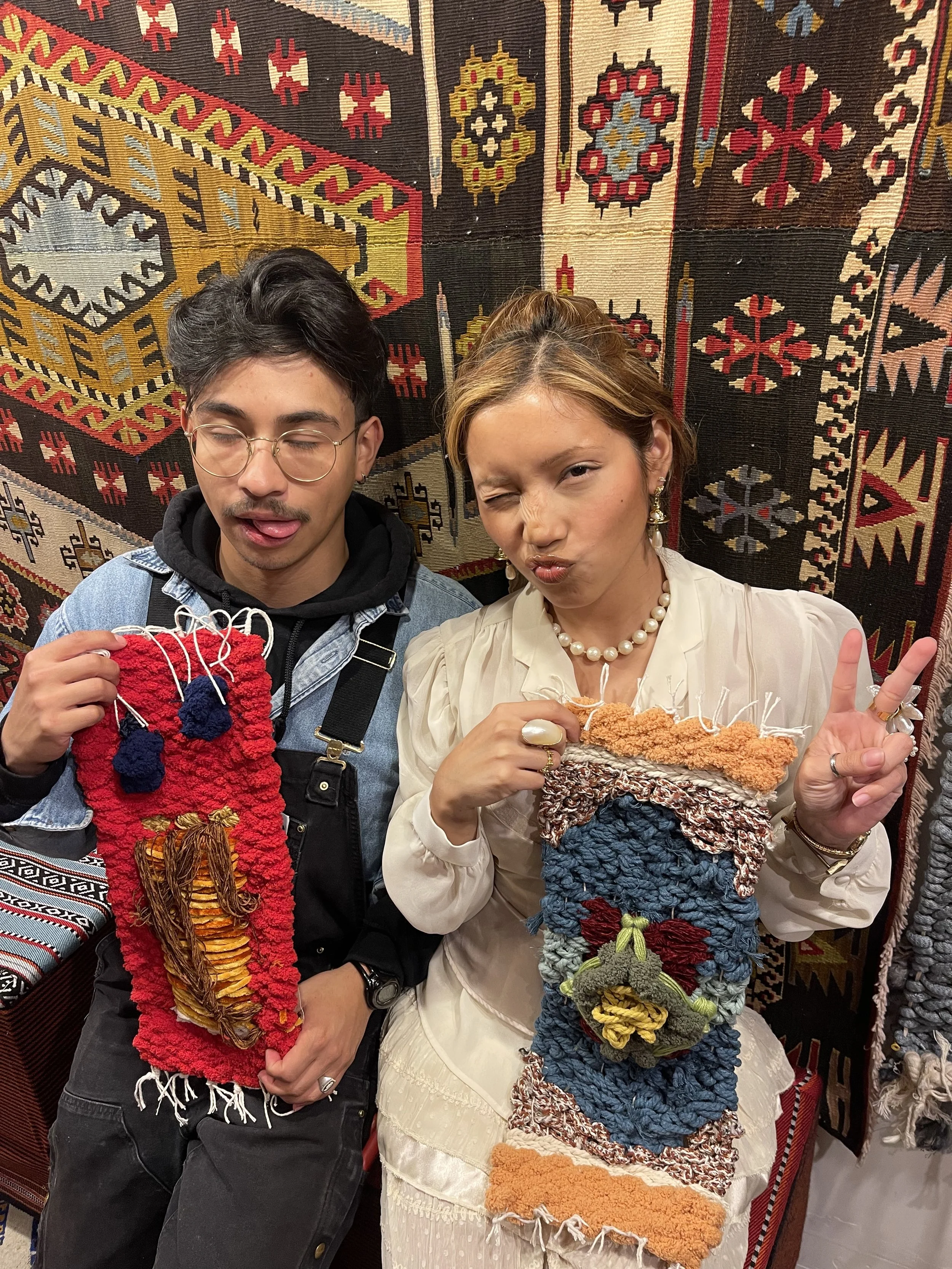 Two people holding colorful, textured woven bags with their faces close to a decorative patterned rug in the background. The person on the left is wearing glasses, a denim jacket, and a black hoodie, and the person on the right is winking, making a p