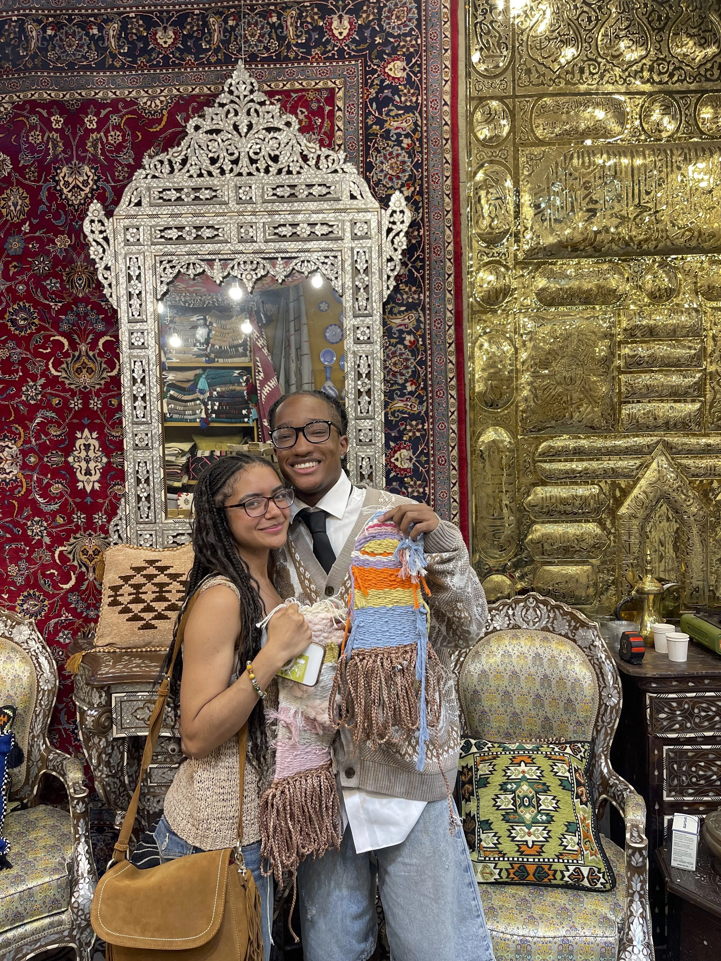 A couple holding their finished hand woven tapestry in front of a handmade mother of pearl mirror from Damascus, Syria, surrounded by an oriental vintage rug and curated textiles inside a New York City weaving studio