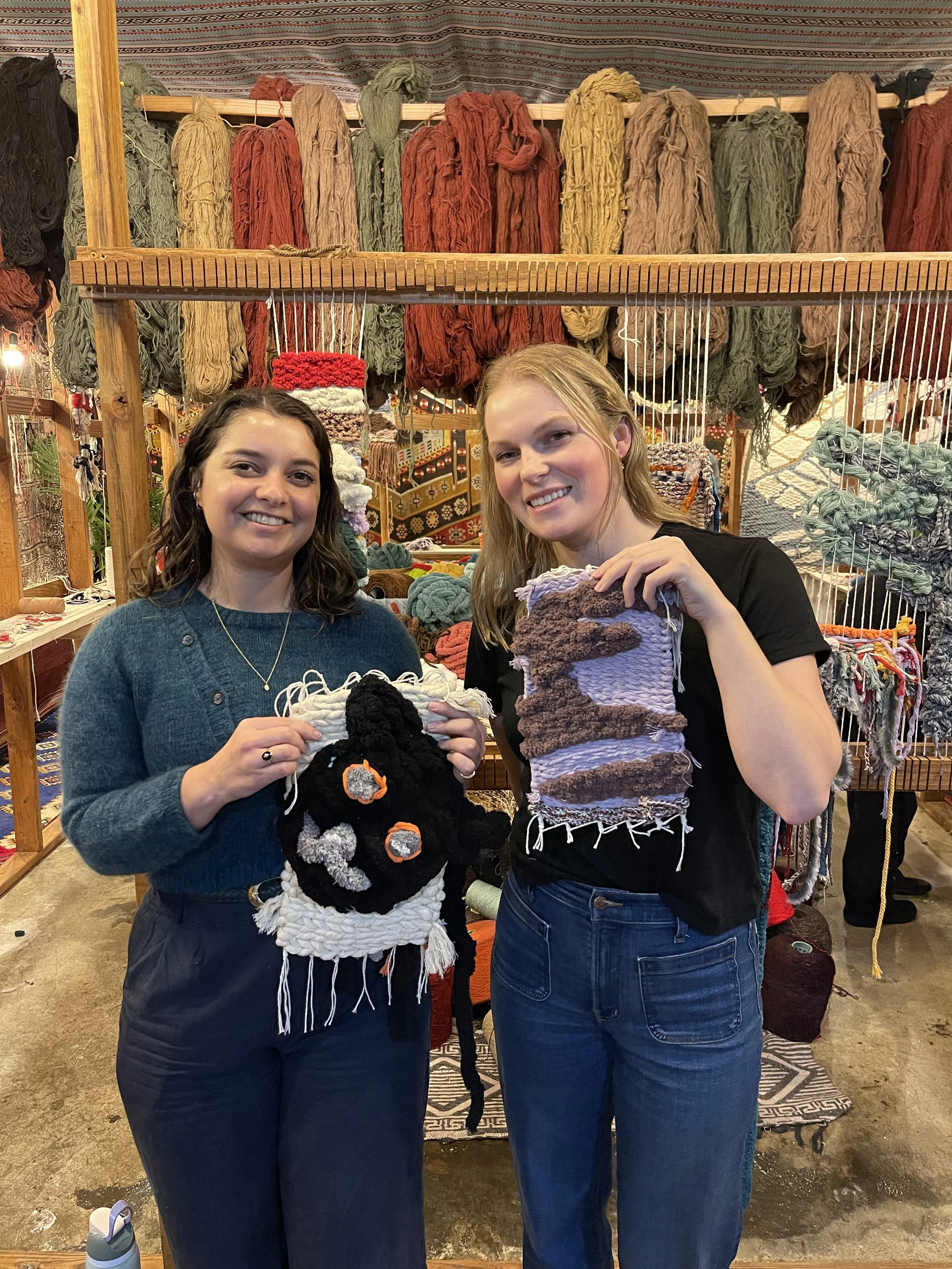 Two women standing in front of a display of colorful yarn at a craft fair, holding handmade woven items.