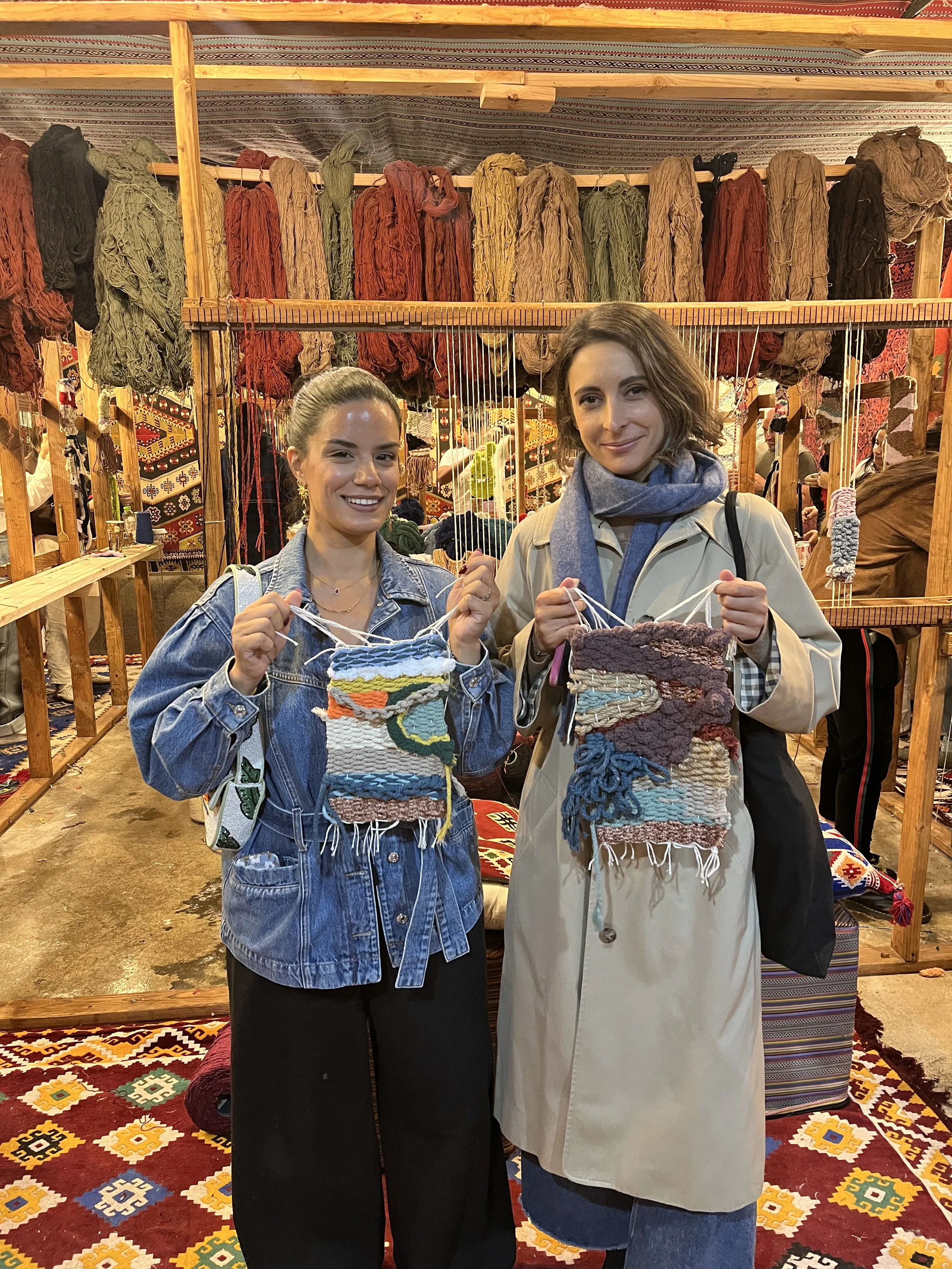 Two women smiling and holding colorful woven rugs at a craft market stall, with yarns hanging behind them.