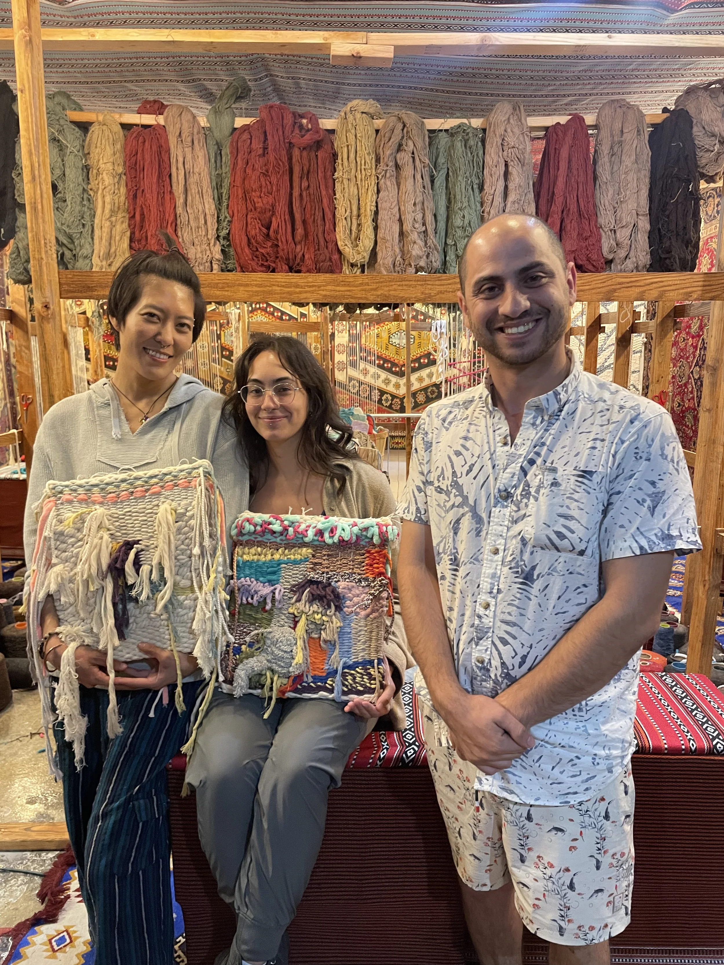 Master Weaver Mahmoud standing with two students proudly displaying their finished tapestry pieces inside the iconic Bedouin-style loom at Creative Minds in Williamsburg, Brooklynsurrounded by handwoven wool and cotton textiles, echoing Syrian.