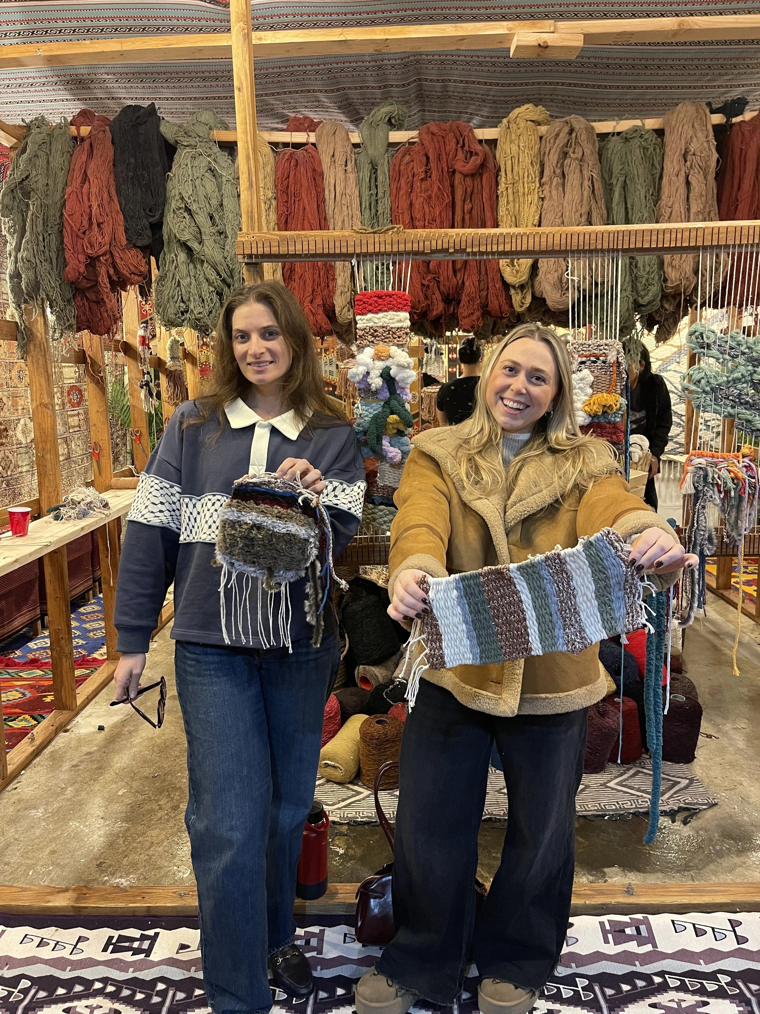 Two women standing in front of a textile and yarn display at a craft fair. They are holding knitted items, with colorful yarn and wool on shelves behind them.