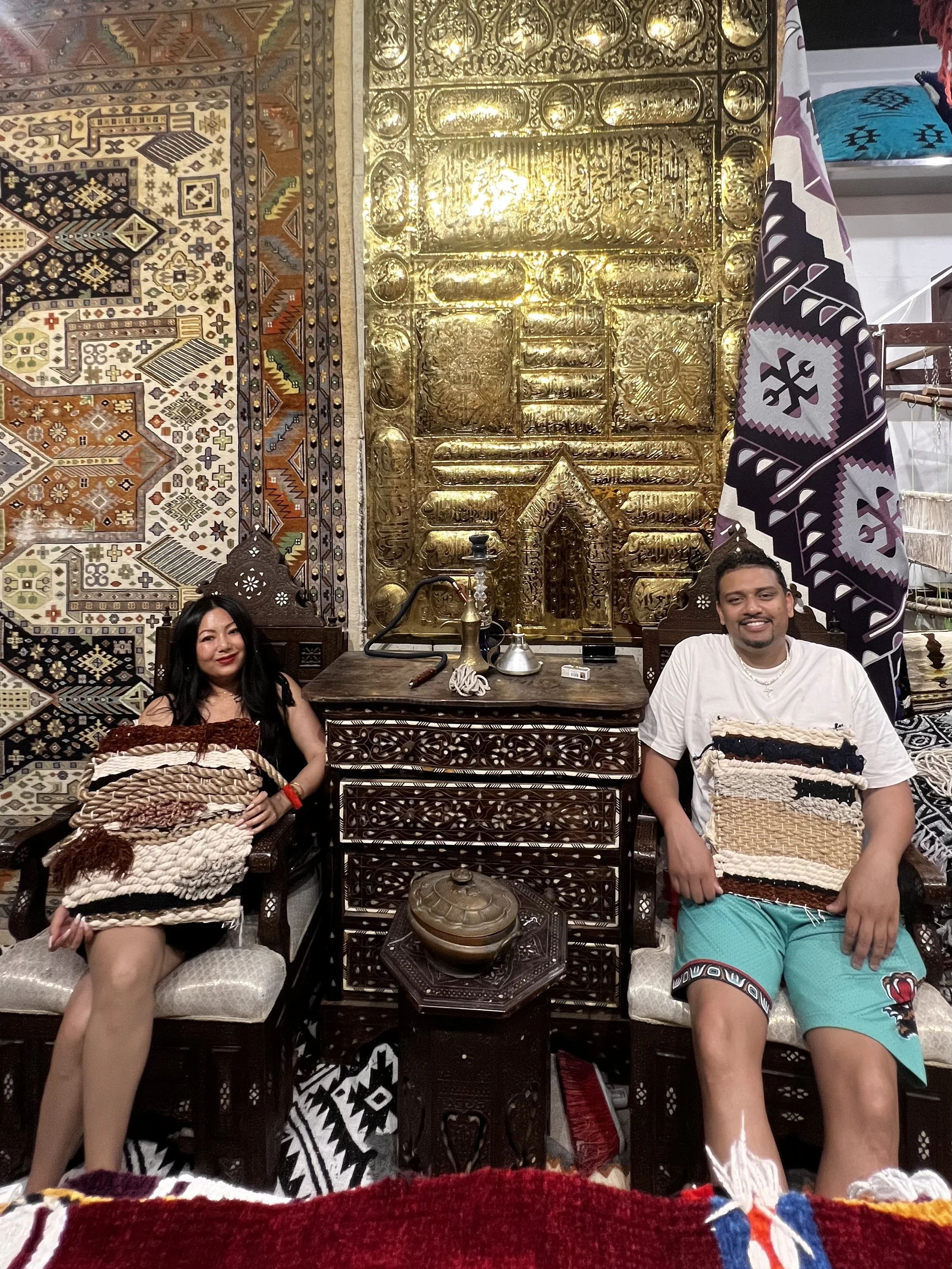 A couple seated on vintage Damascus furniture at Creative Minds in Williamsburg, Brooklyn, proudly posing with their handmade tapestries created during the weaving class blending Syrian heritage with contemporary artistry.