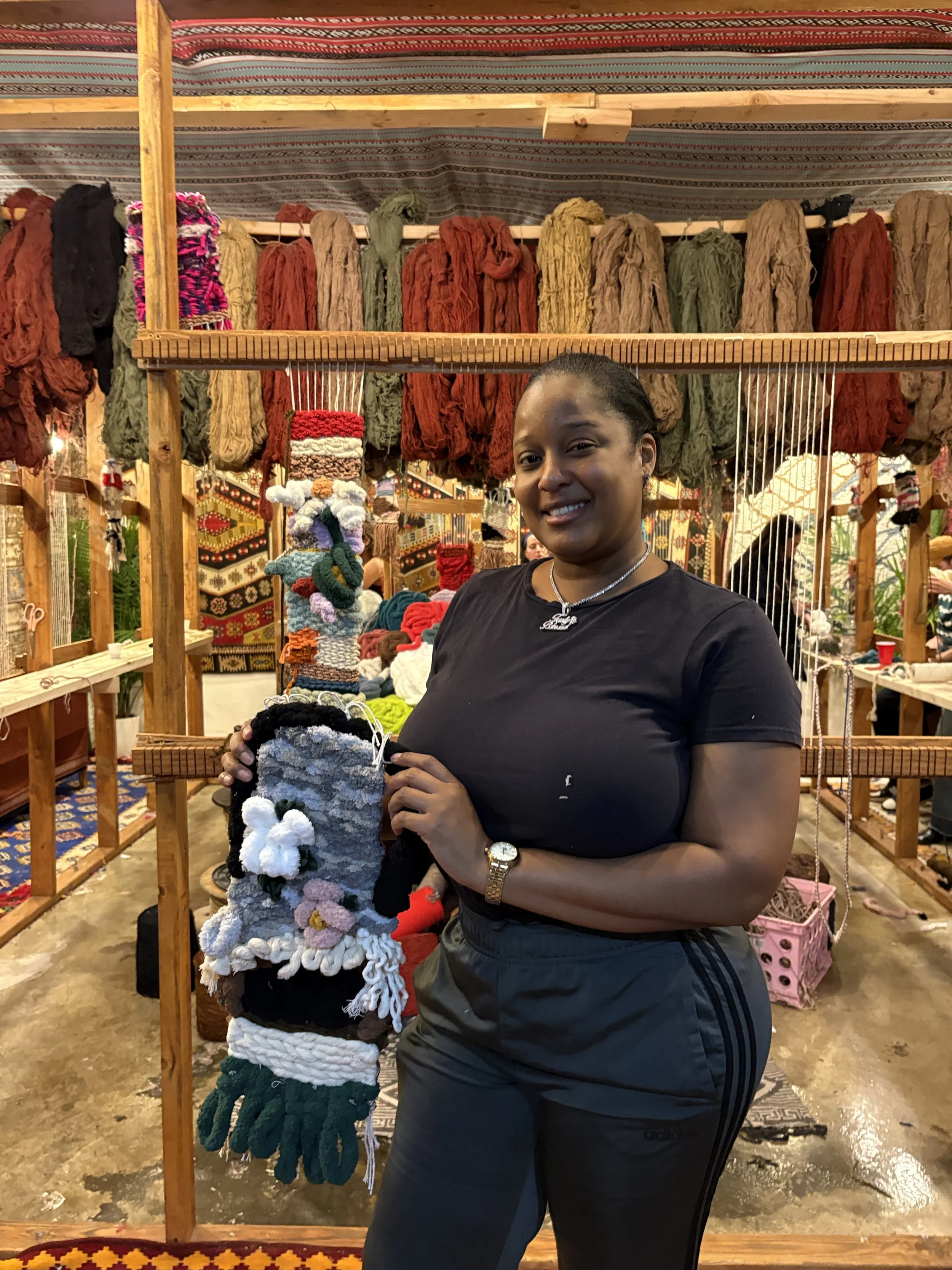 A woman is standing in front of a display of colorful woven and handmade textiles, holding a decorated woven item with various colors and textures in a market or craft fair setting.