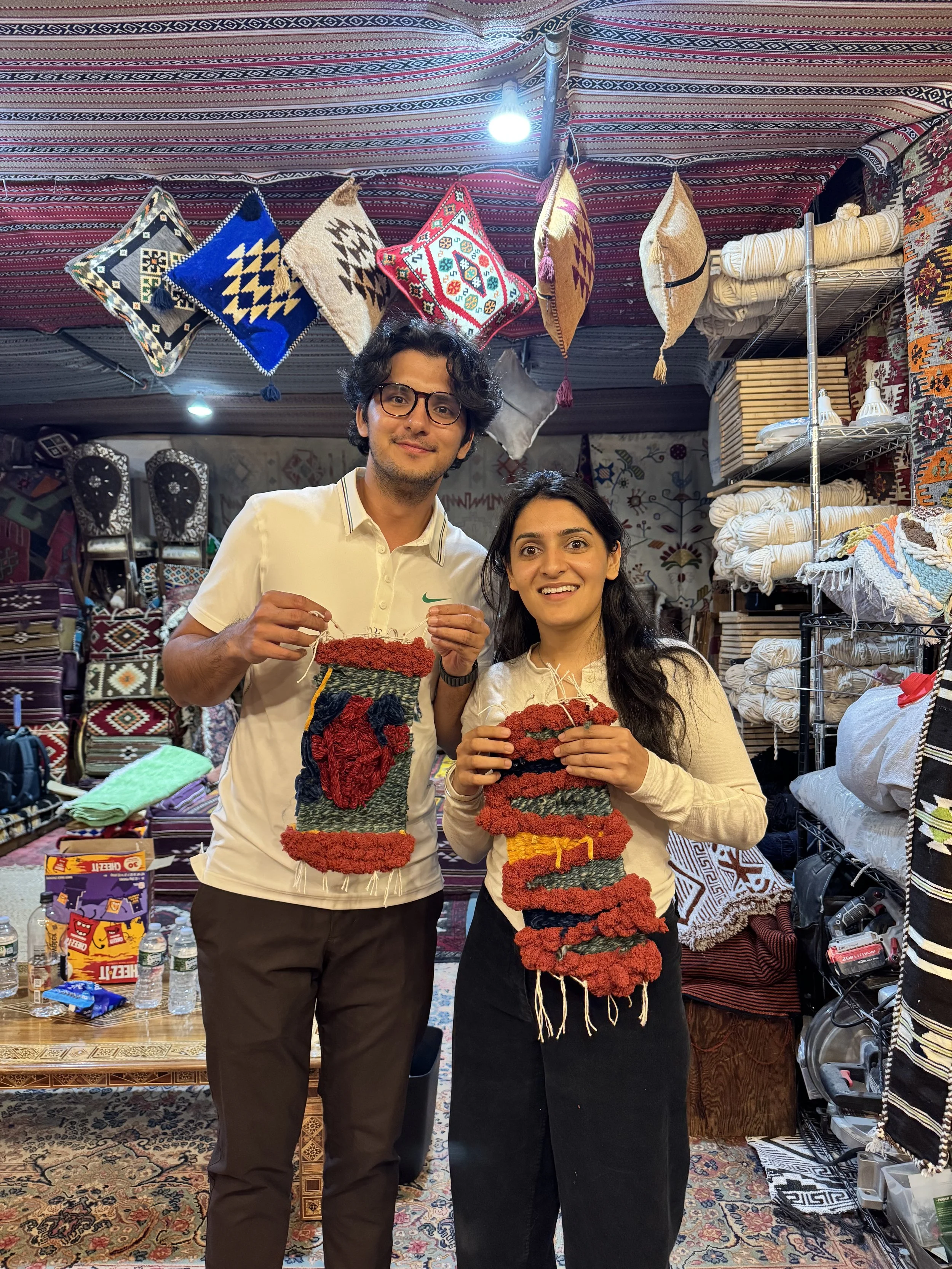 Two people, a man and a woman, standing inside a textiles shop, holding colorful woven wall hangings or rugs.