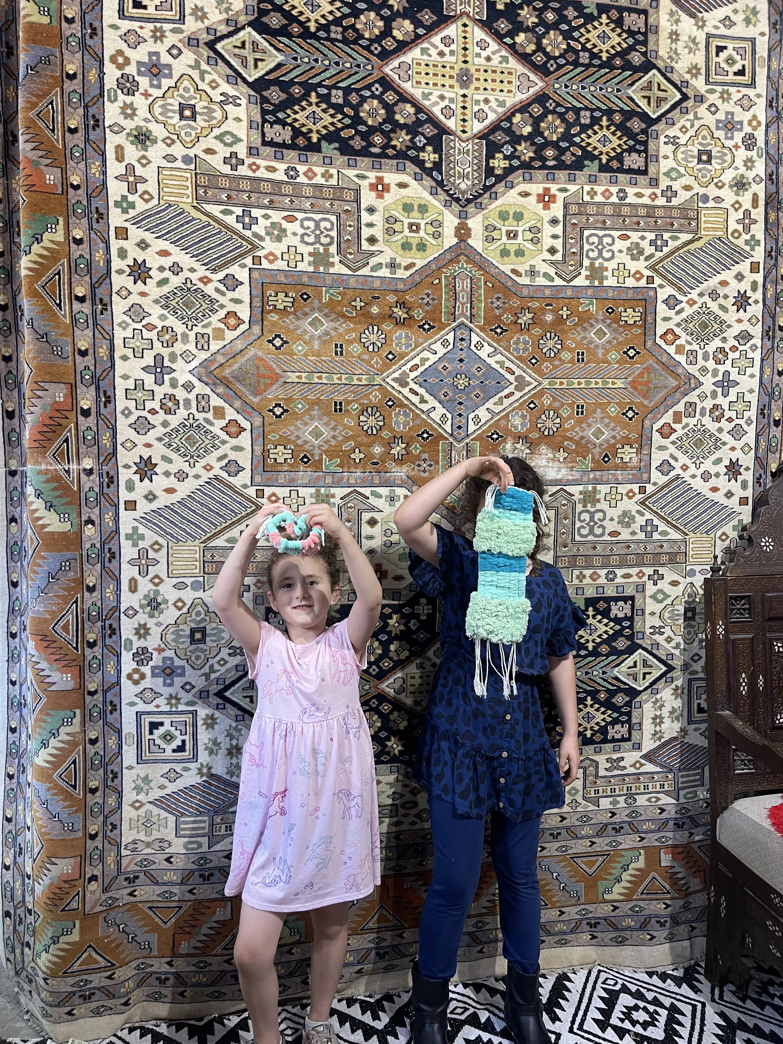 Two children proudly displaying their handmade tapestries created at the iconic loom inside Creative Minds in Williamsburg, Brooklyn standing next to a vintage Oriental rug, celebrating creativity and cultural tradition through weaving.