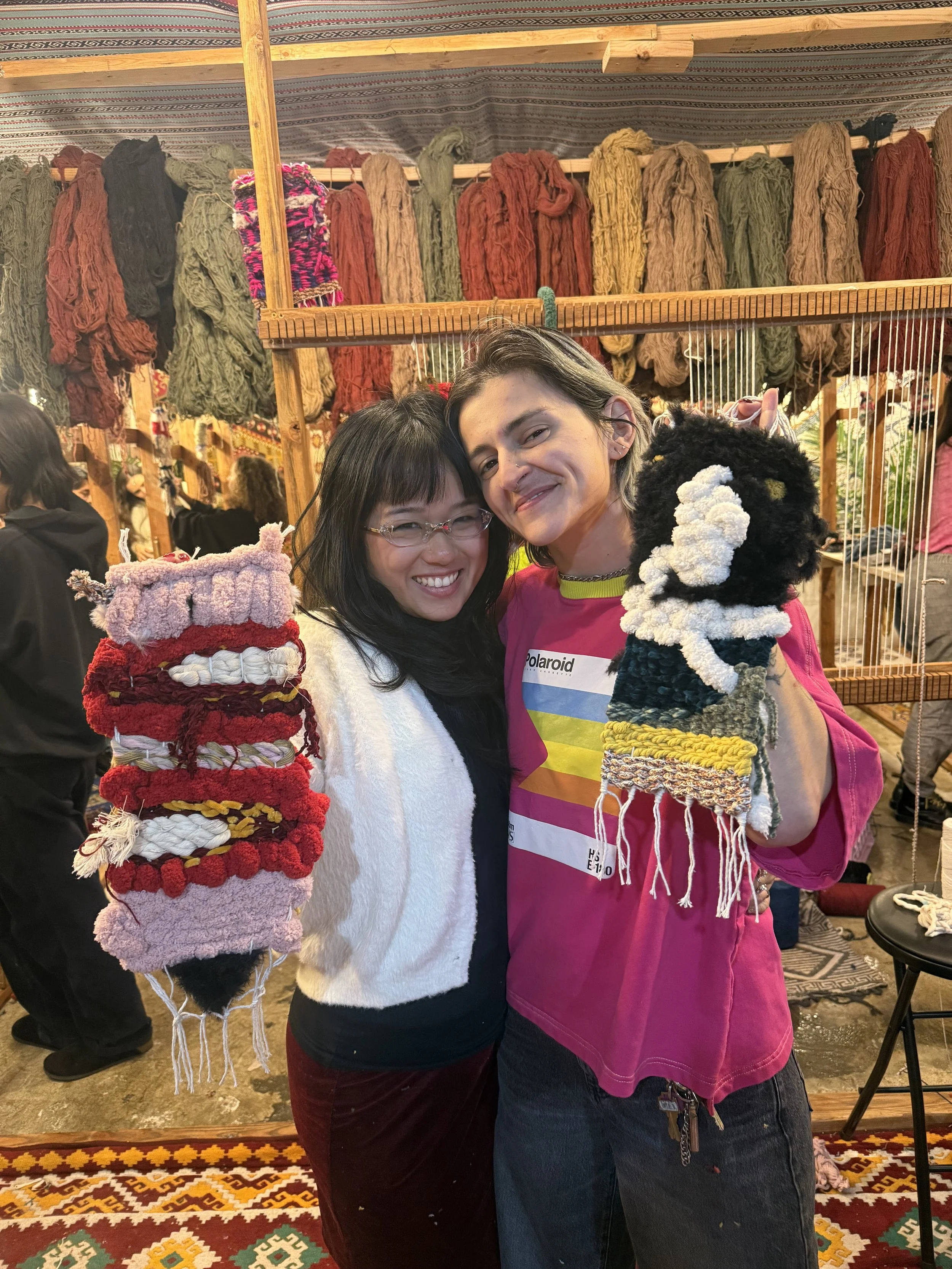 Two women smiling and holding handmade woven puppets, standing in front of a display of colorful yarn in a craft stall.