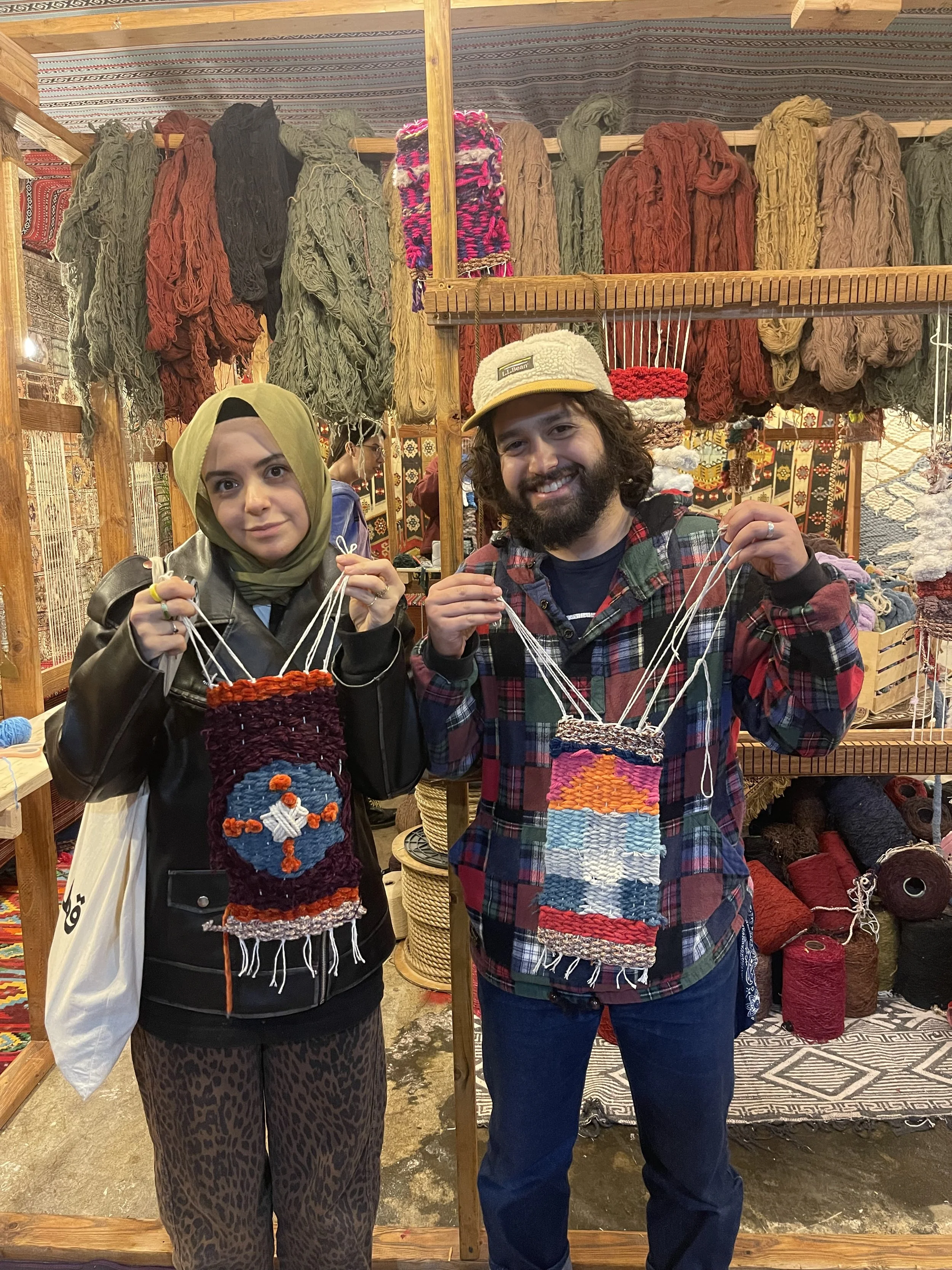 Two people holding woven textile art pieces inside a craft stall with colorful yarns hanging in the background.