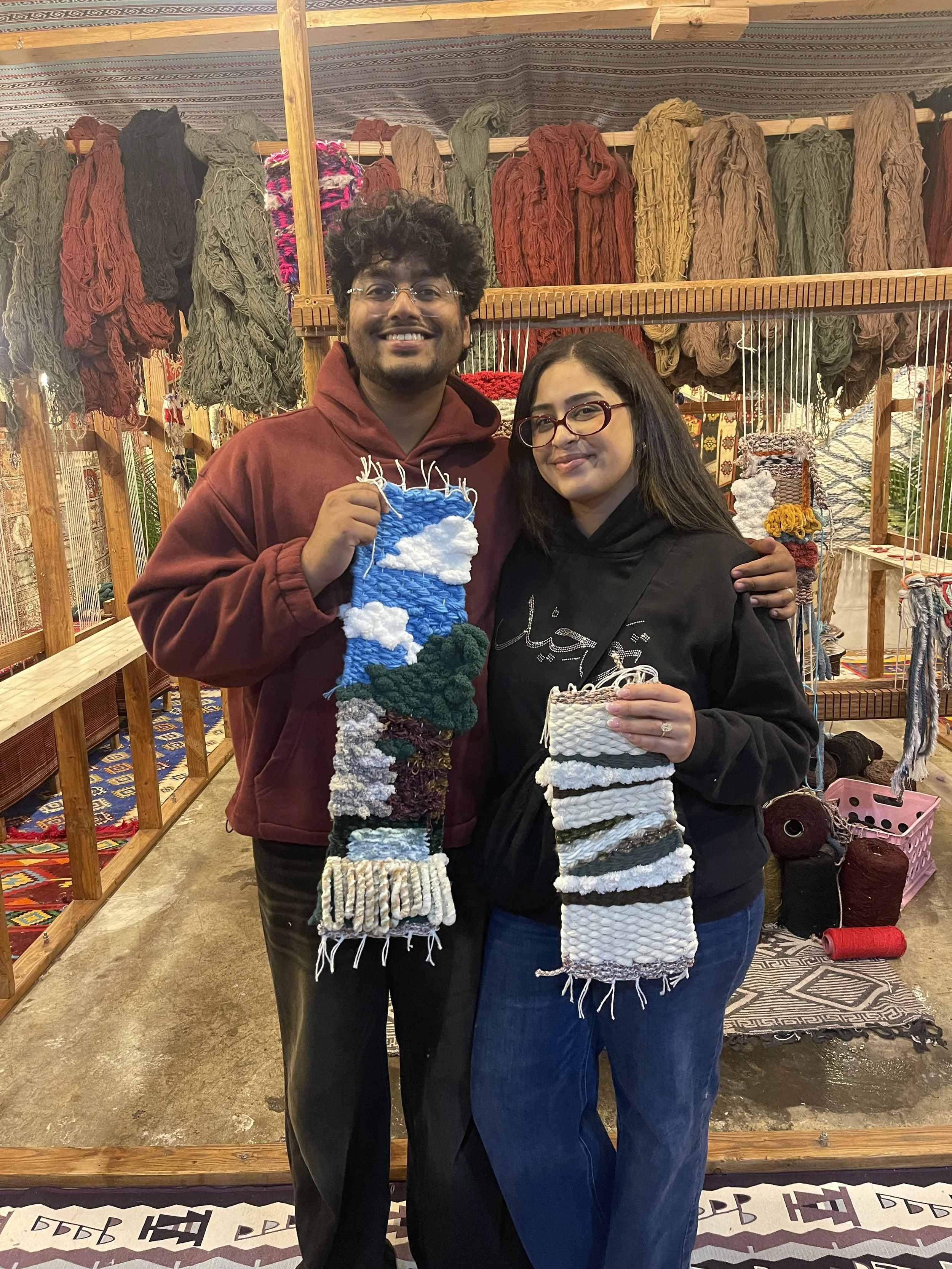 A man and woman smiling inside a handcrafted textile shop, holding colorful woven textile pieces. The background shows shelves with multicolored yarns and traditional woven fabric displays.