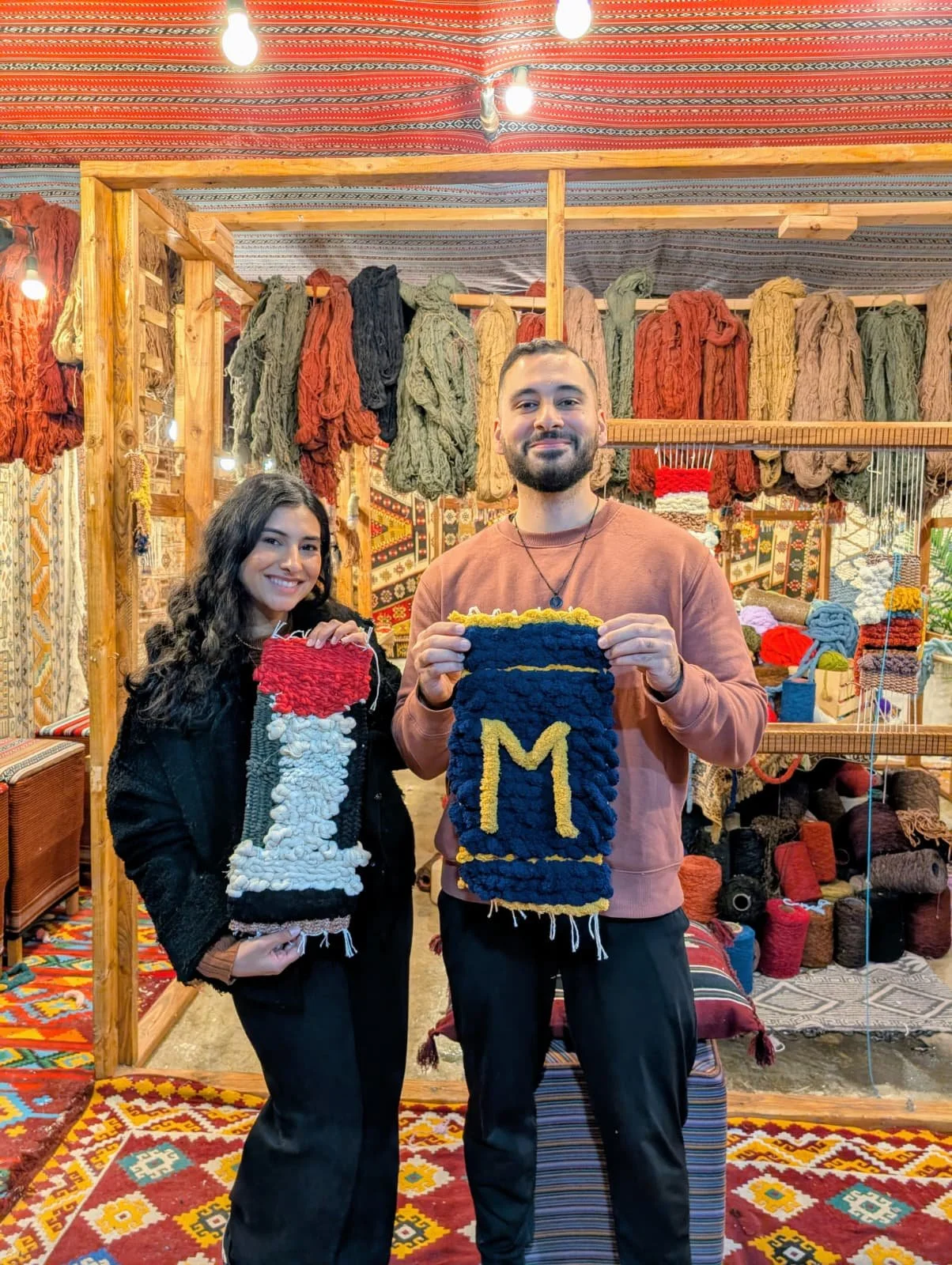 Two people smiling and holding handmade woven items in a colorful craft booth with yarn and textiles in the background.