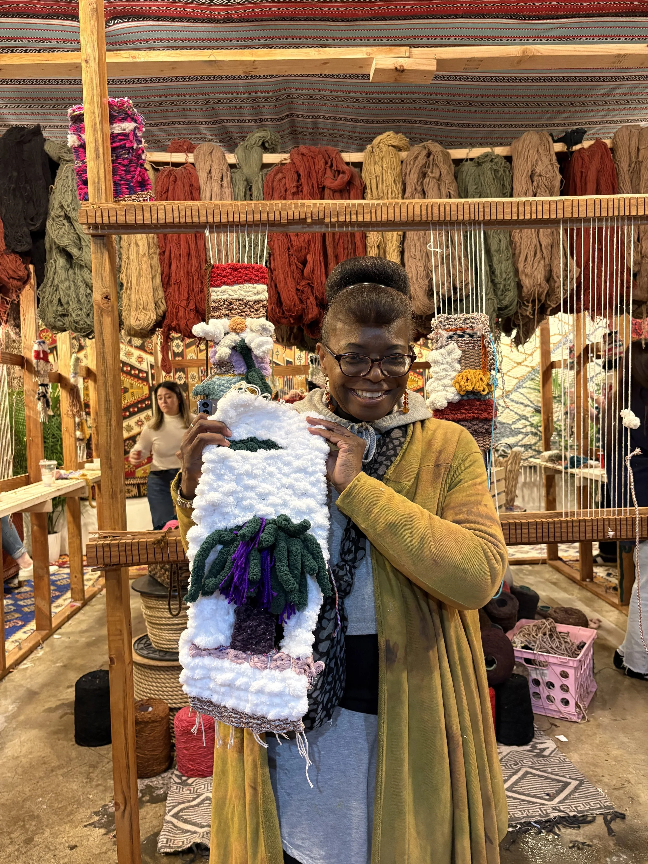 A woman standing in a craft market holding a small, handmade yarn bag with a face of a character with green hair and purple accents. Behind her are racks of colorful yarn and other handmade items, with other shoppers visible in the background.