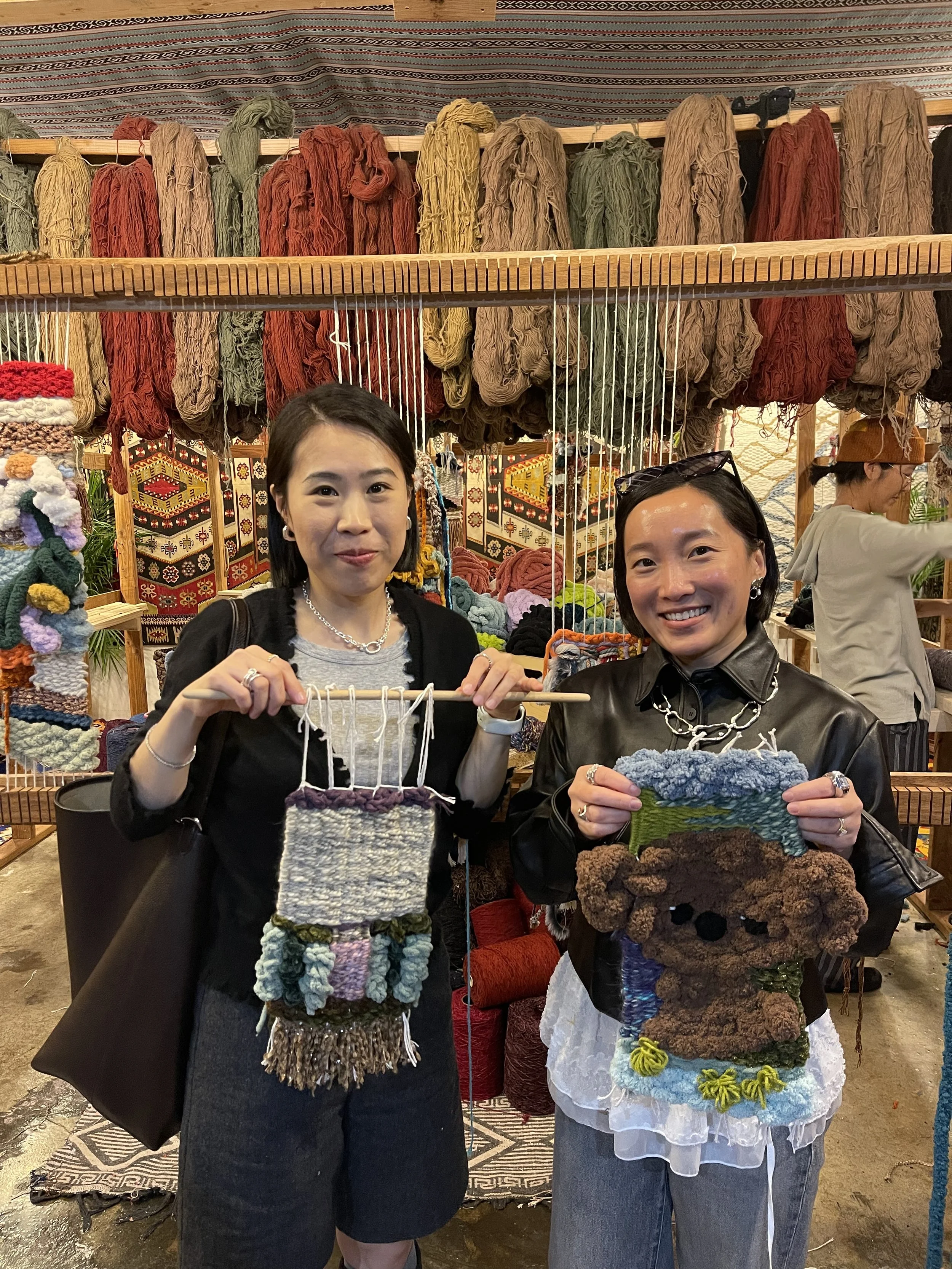 Two women at a craft fair holding small woven or felted art pieces with a display of colorful yarn in the background.