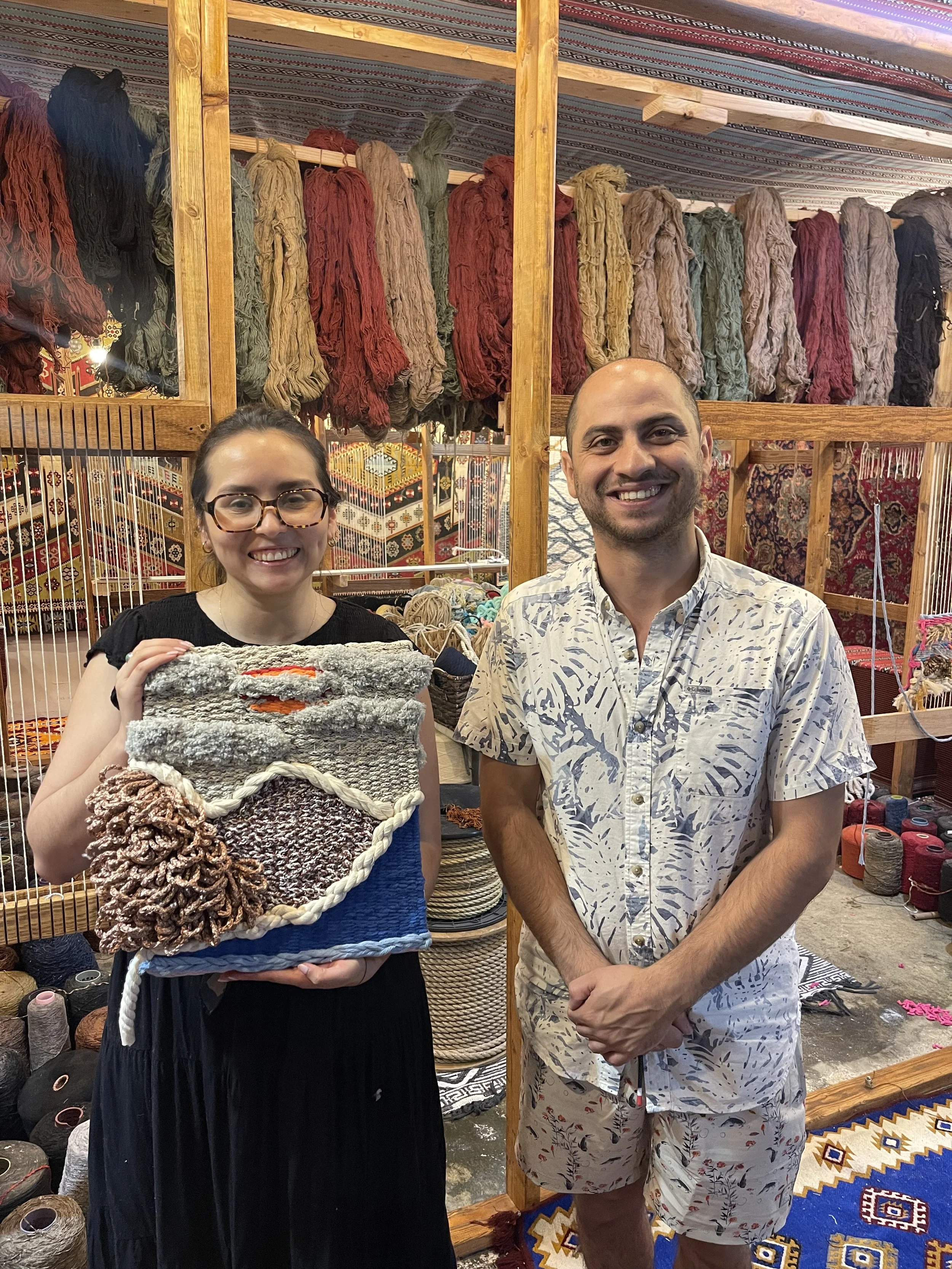 Master Weaver Mahmoud standing beside the iconic loom at Creative Minds in Williamsburg, Brooklyn, with a customer proudly holding their finished tapestry celebrating the fusion of traditional Syrian weaving and personal creativity