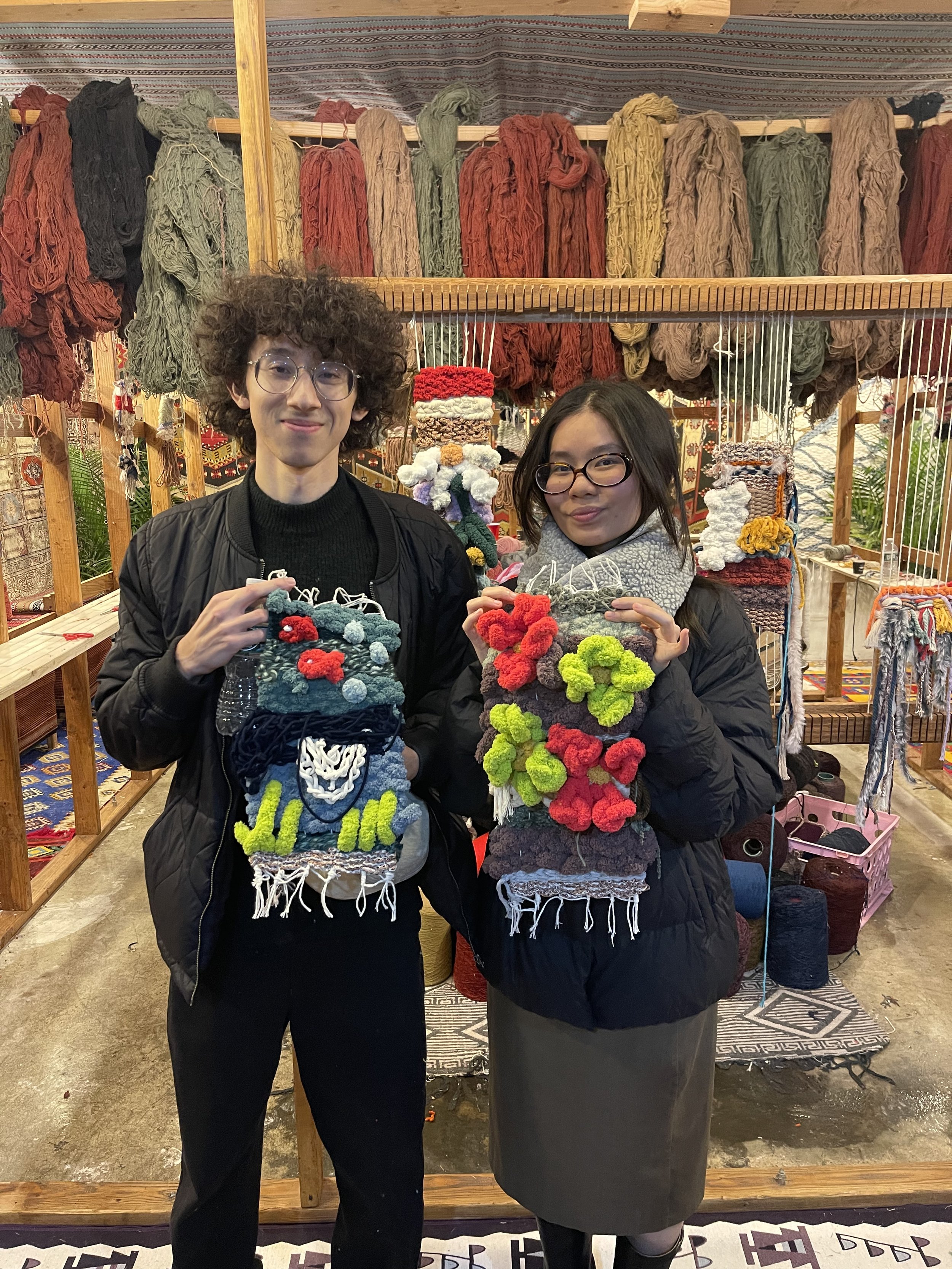 Two individuals, a young man and woman, holding colorful knitted or crocheted bags with pom-poms, standing in front of a display of yarn and textile products at a craft market.