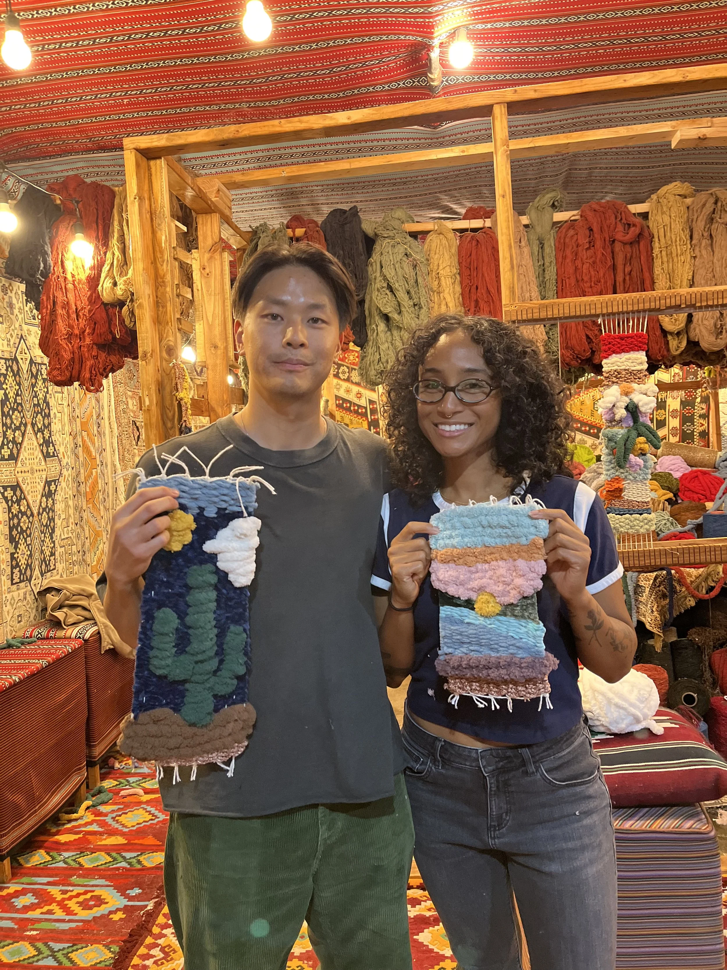 Two smiling people holding small woven fiber art pieces inside a colorful textile shop. The shop has patterned hanging fabrics and yarns on shelves in the background.