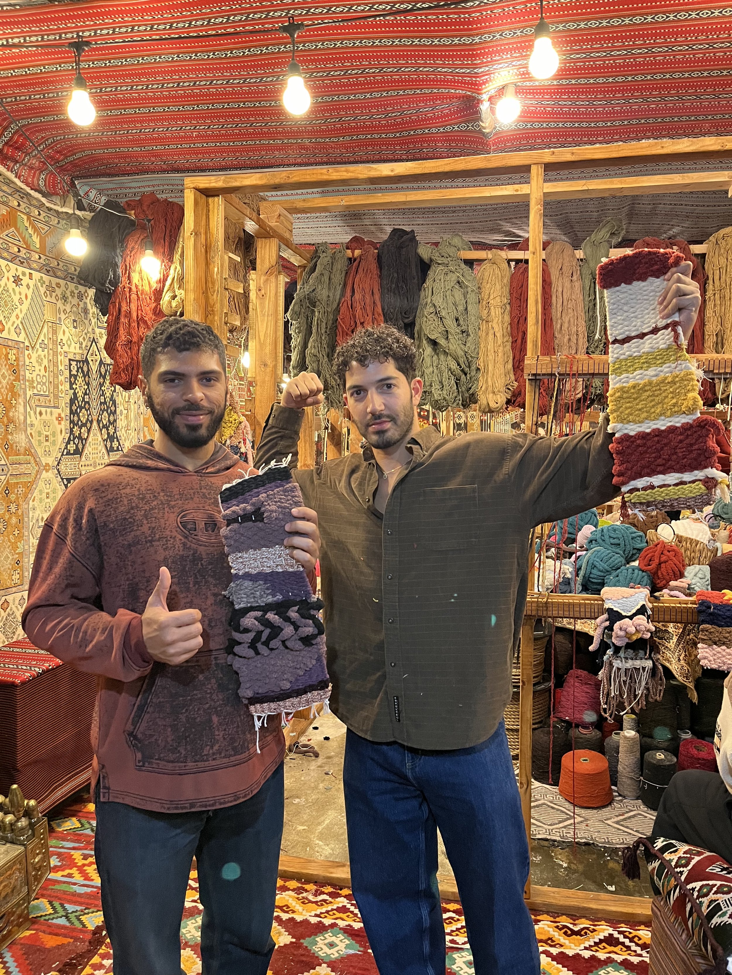 Two men inside a colorful textile shop displaying knitted socks and scarves, with vibrant woven rugs and yarns in the background.