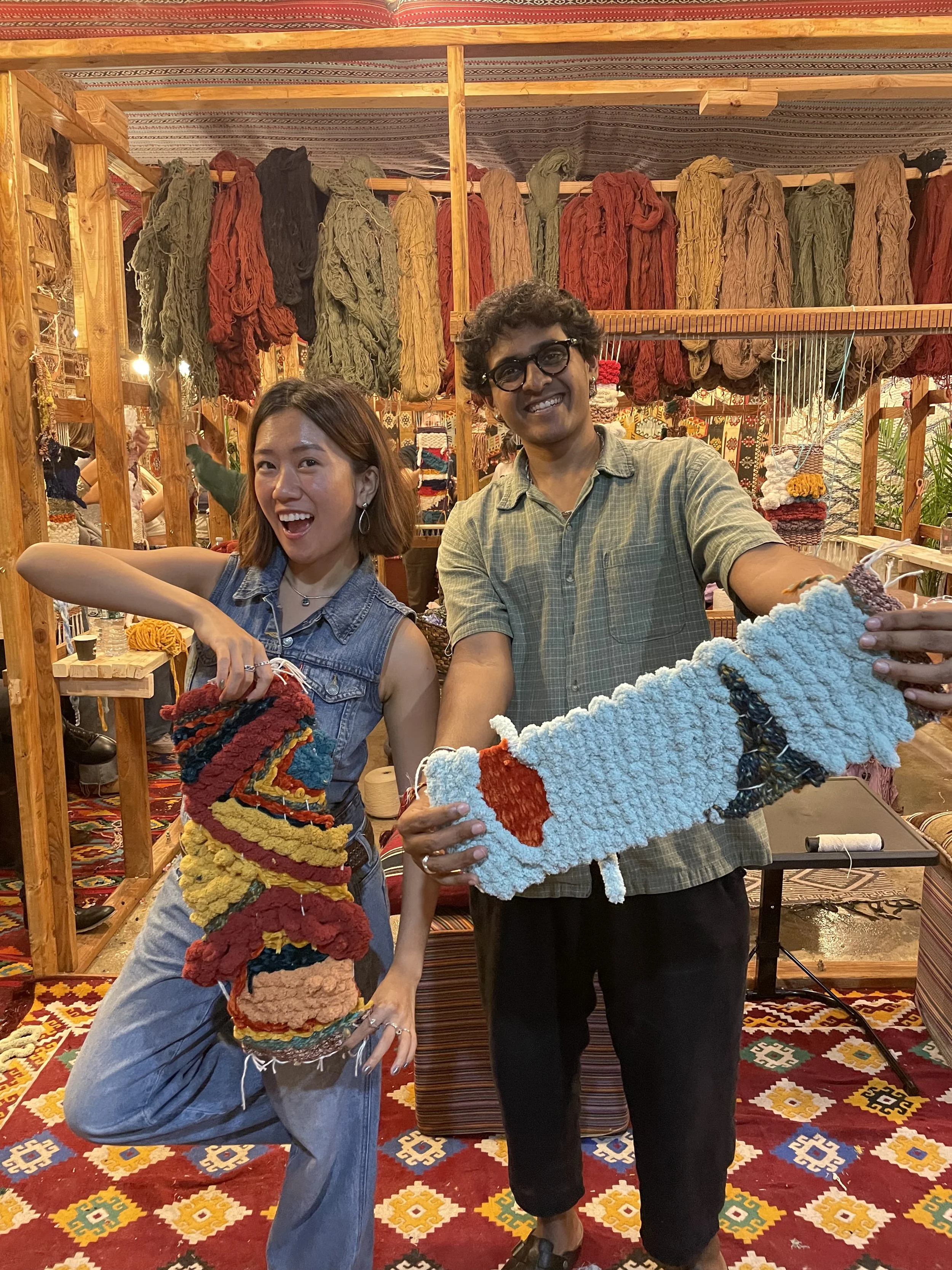 Two people smiling and holding colorful woven or knitted crafts inside a vibrant, decorated craft shop featuring hanging yarns and patterned rugs.