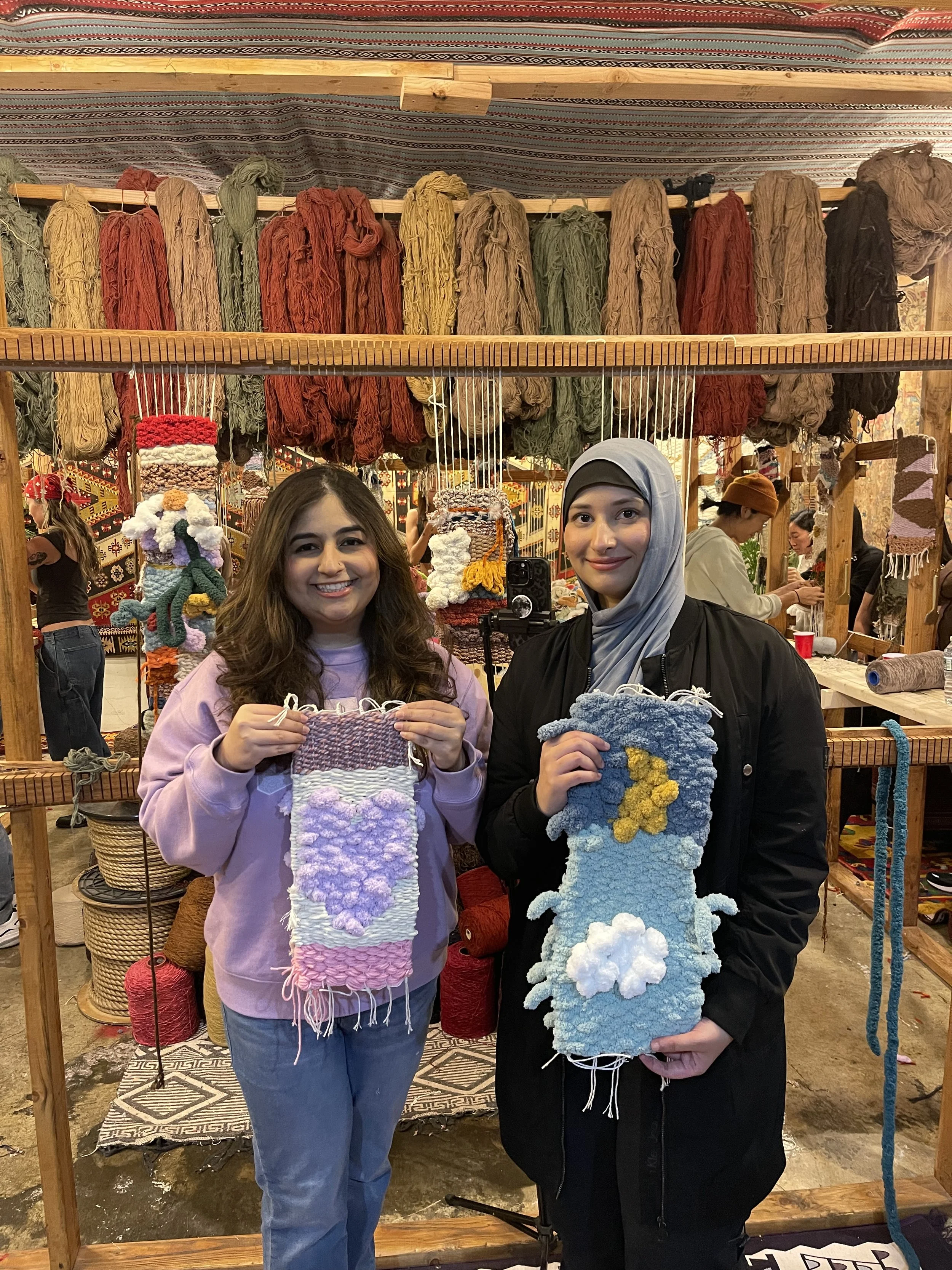 Two women are standing in front of a display of colorful yarn at a craft fair, each holding a small, textured crochet piece with cloud and rainbow motifs.