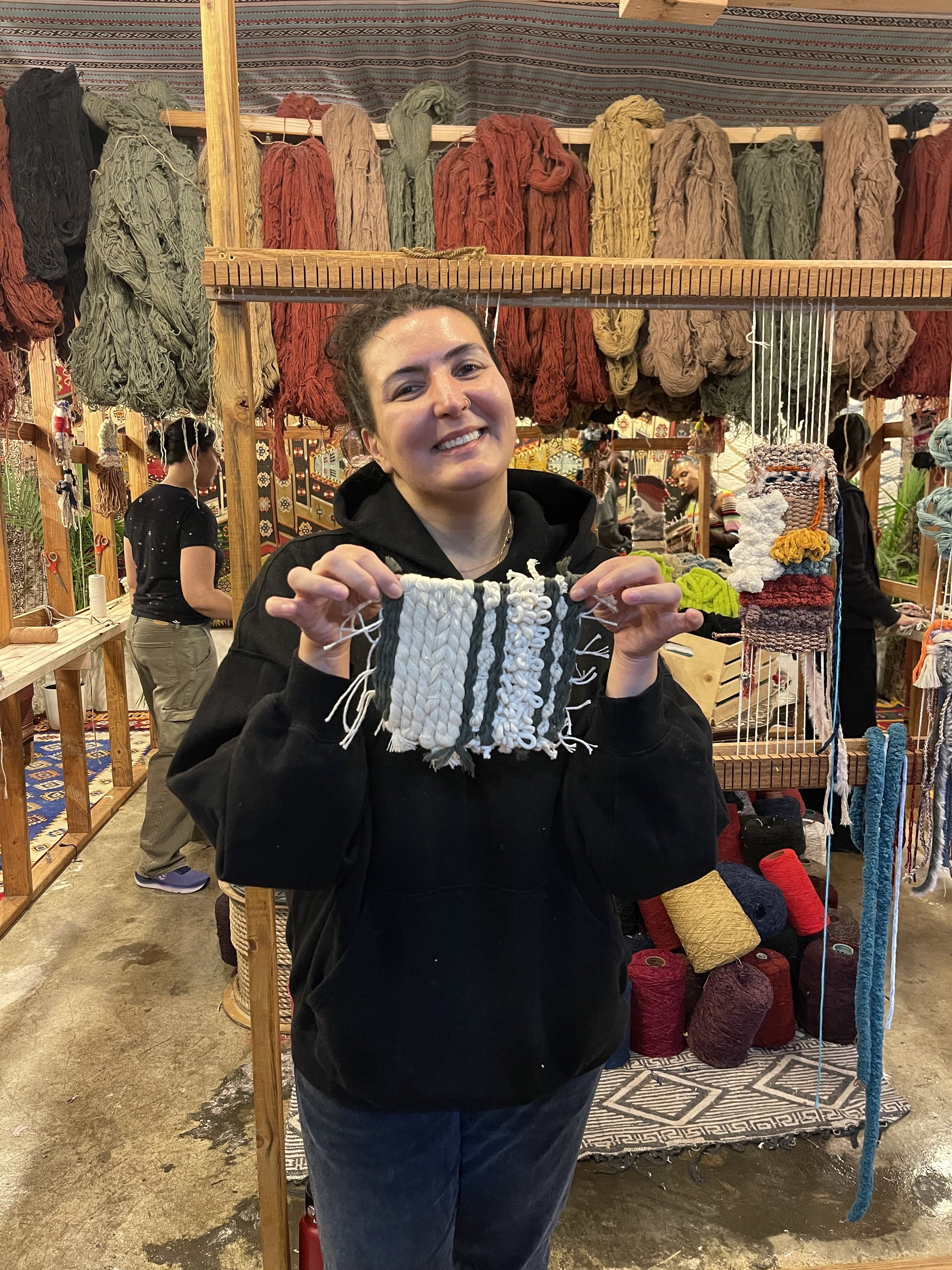 A woman is smiling and holding a piece of woven textile with black and white stripes, standing in a craft market with colorful yarns and woven items displayed behind her.