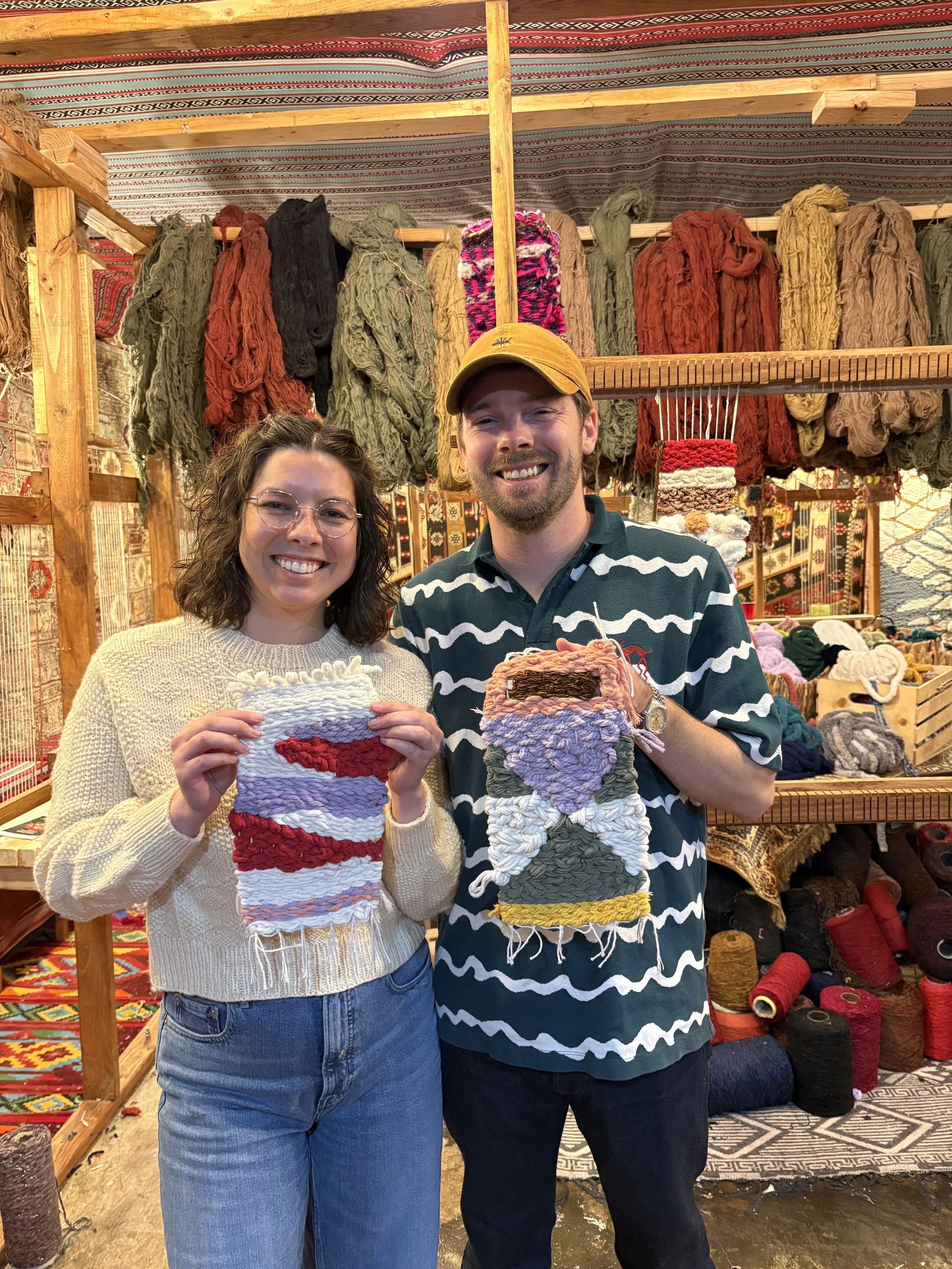 Two people smiling and holding handmade woven items inside a craft stall with colorful yarns and woven textiles displayed on shelves and walls.