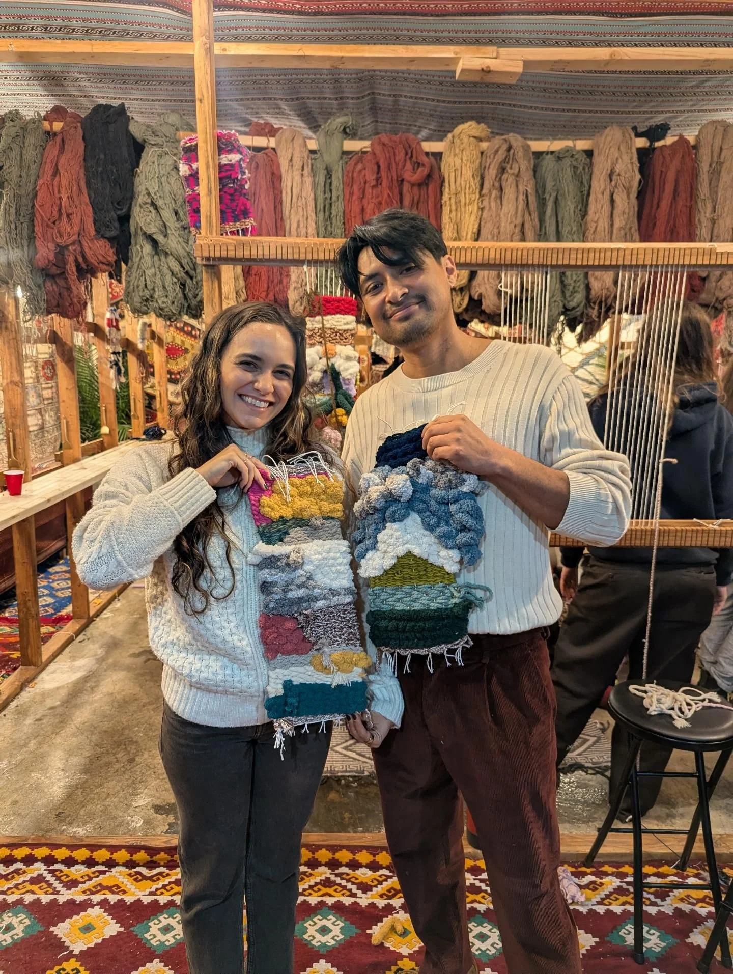 Two people smiling and holding colorful woven or knitted items in a craft booth with various yarns hanging in the background.