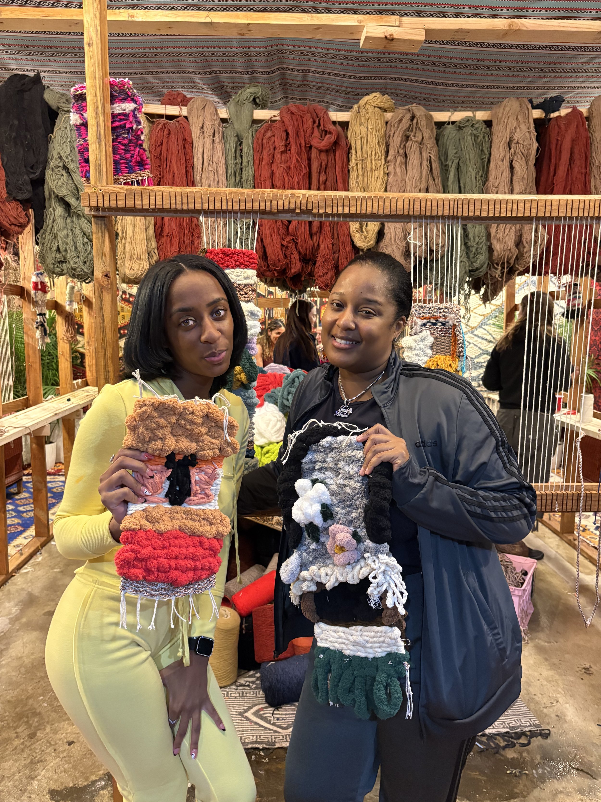 Two women standing in front of a colorful yarn display, holding knitted or crocheted items, at a craft fair or market.