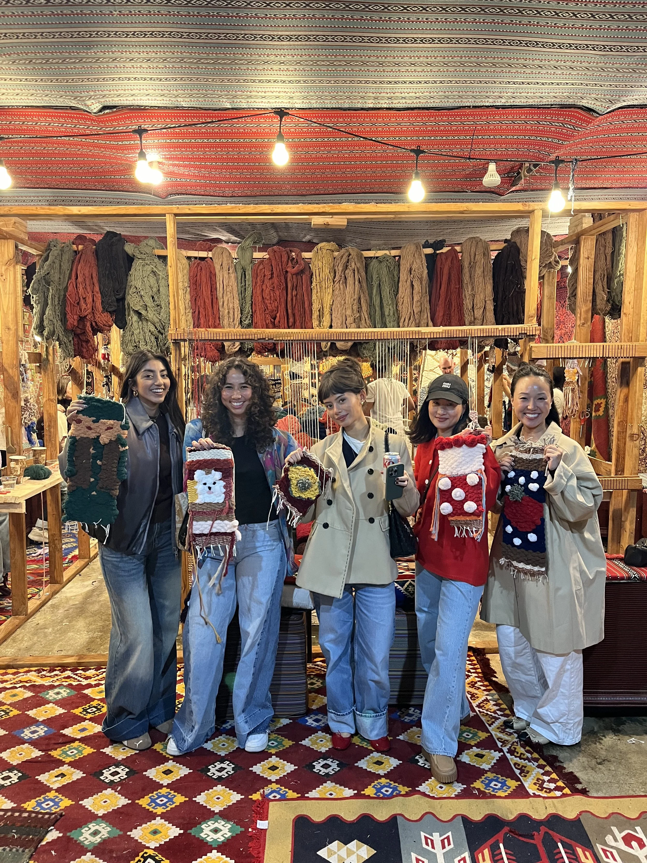 Five women standing in a craft booth with handmade woven socks, smiling and holding their creations. The booth displays colorful yarns and woven textiles in the background.