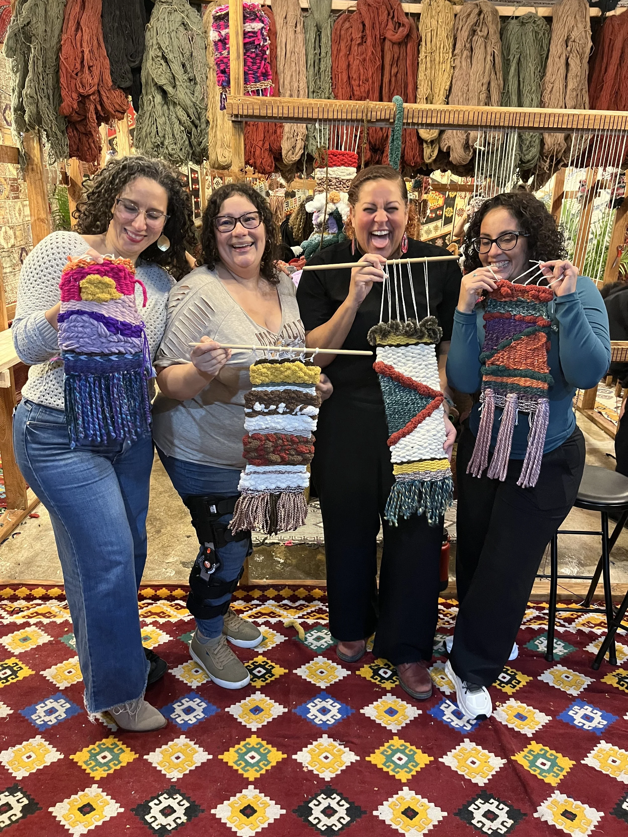 Four women standing inside a yarn store, holding colorful woven wall hangings and smiling at the camera.