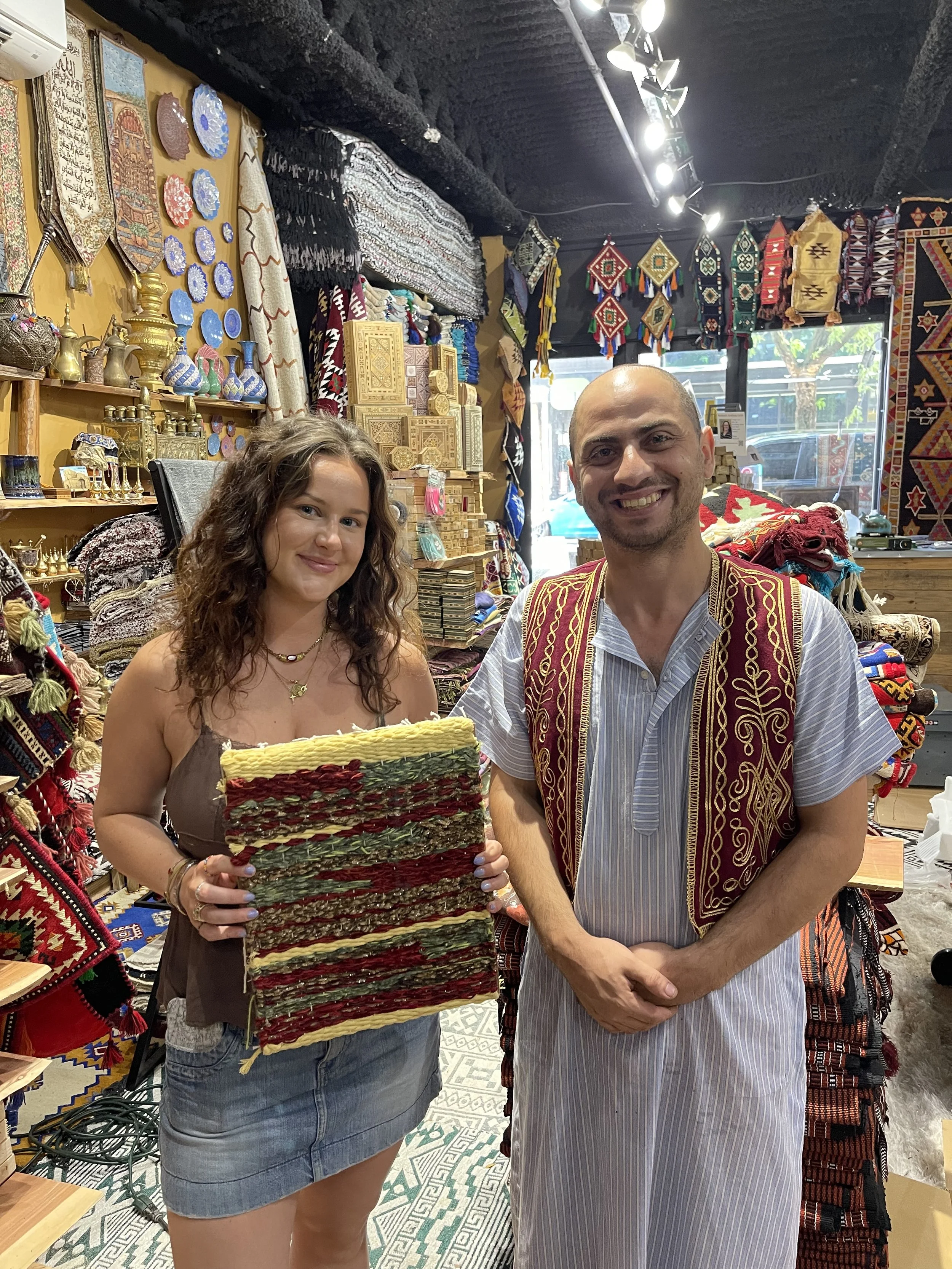 Master Weaver Mahmoud standing beside his first workshop customer in the upstairs retail area of Creative Minds in Williamsburg, Brooklyn proudly showcasing the tapestry they created together in the weaving studio.