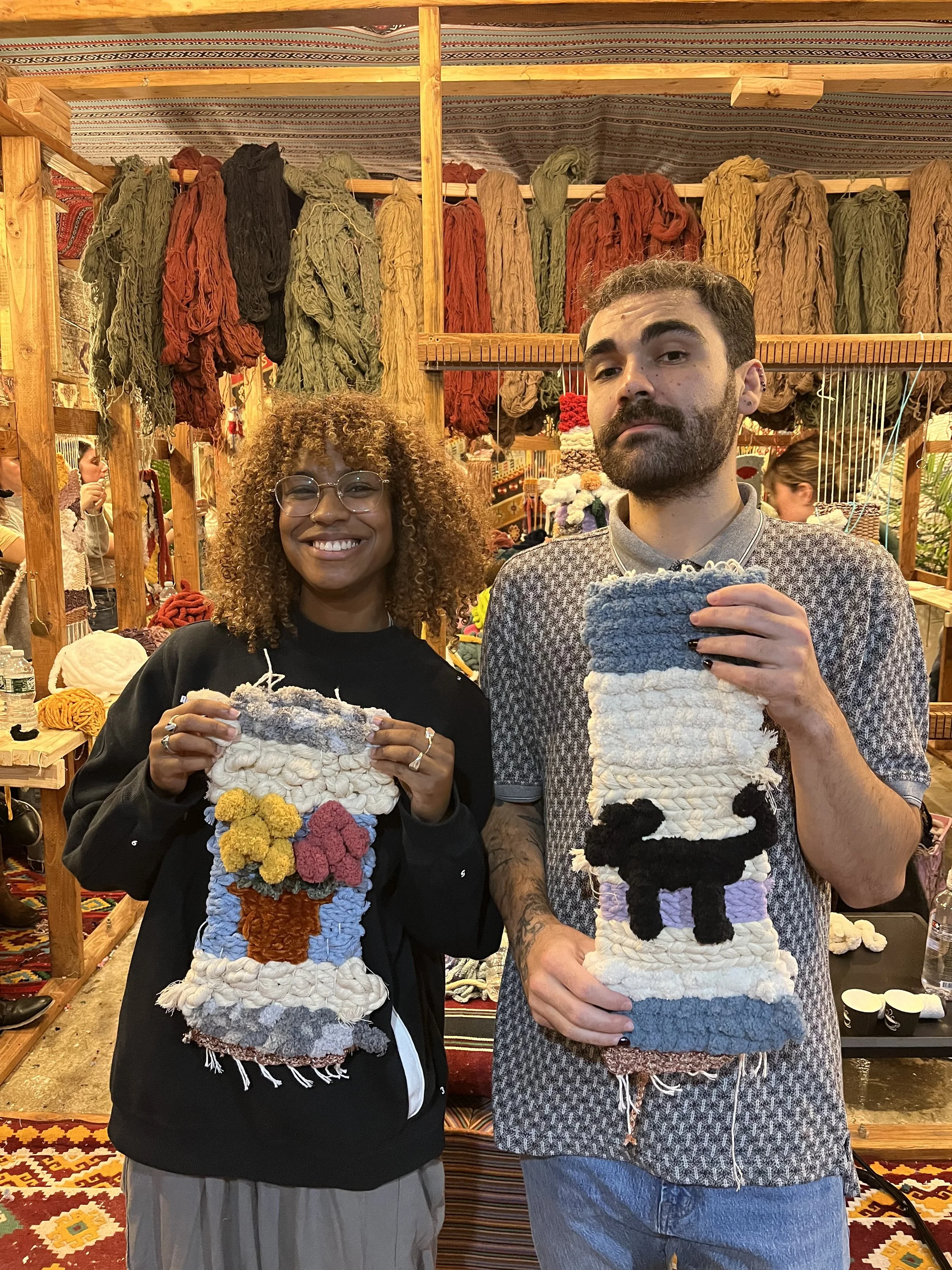A woman and a man standing indoors at a craft market, holding up colorful woven wall hangings with a floral design and a dog silhouette, respectively. Background includes yarn and craft supplies, with woven fabrics hanging above.