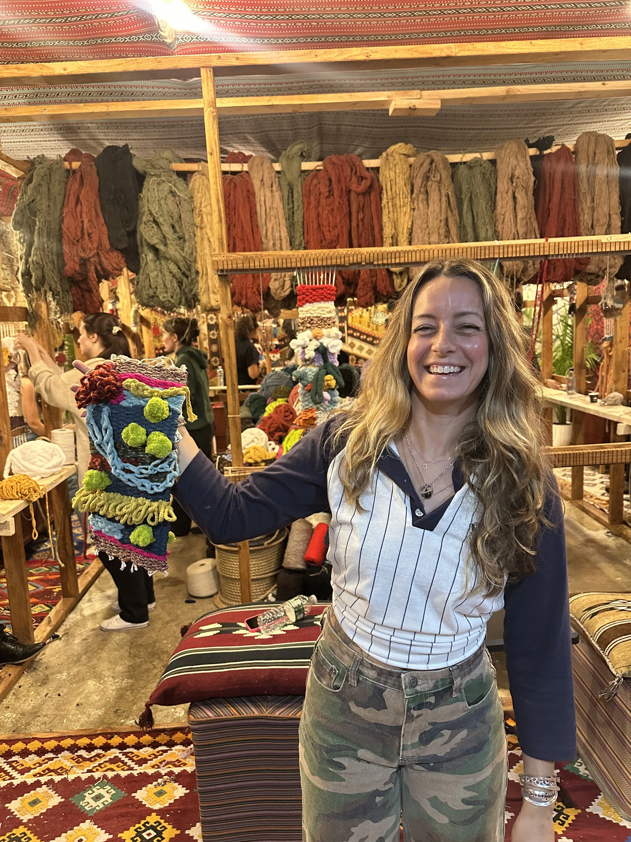 A smiling woman in casual camouflage pants and a striped shirt, holding a colorful, handmade fiber art piece in a craft market with yarn and textile displays in the background.