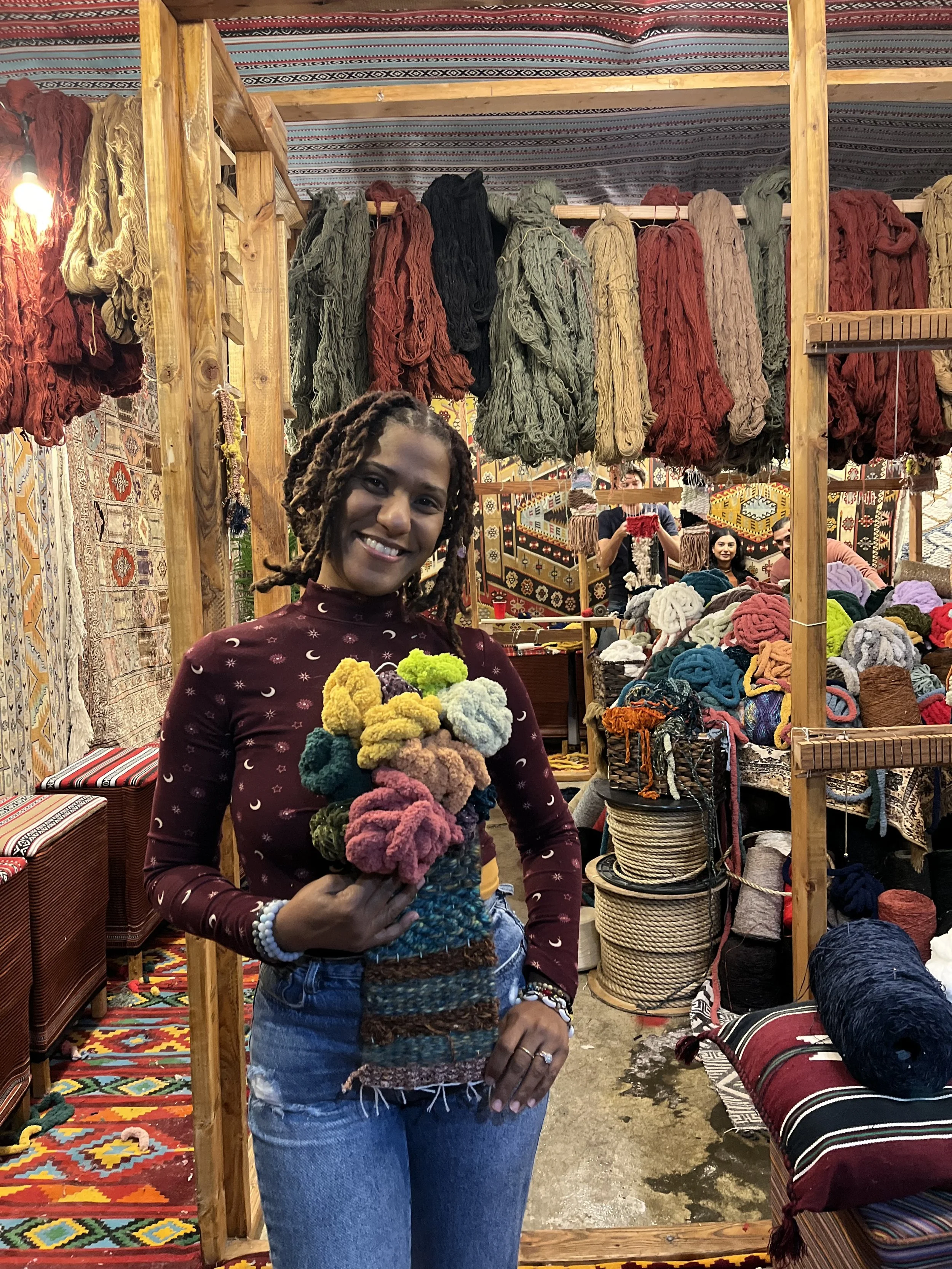 Woman smiling and holding a bunch of colorful yarn in a market stall with hanging yarn, woven rugs, and various colorful textile supplies.