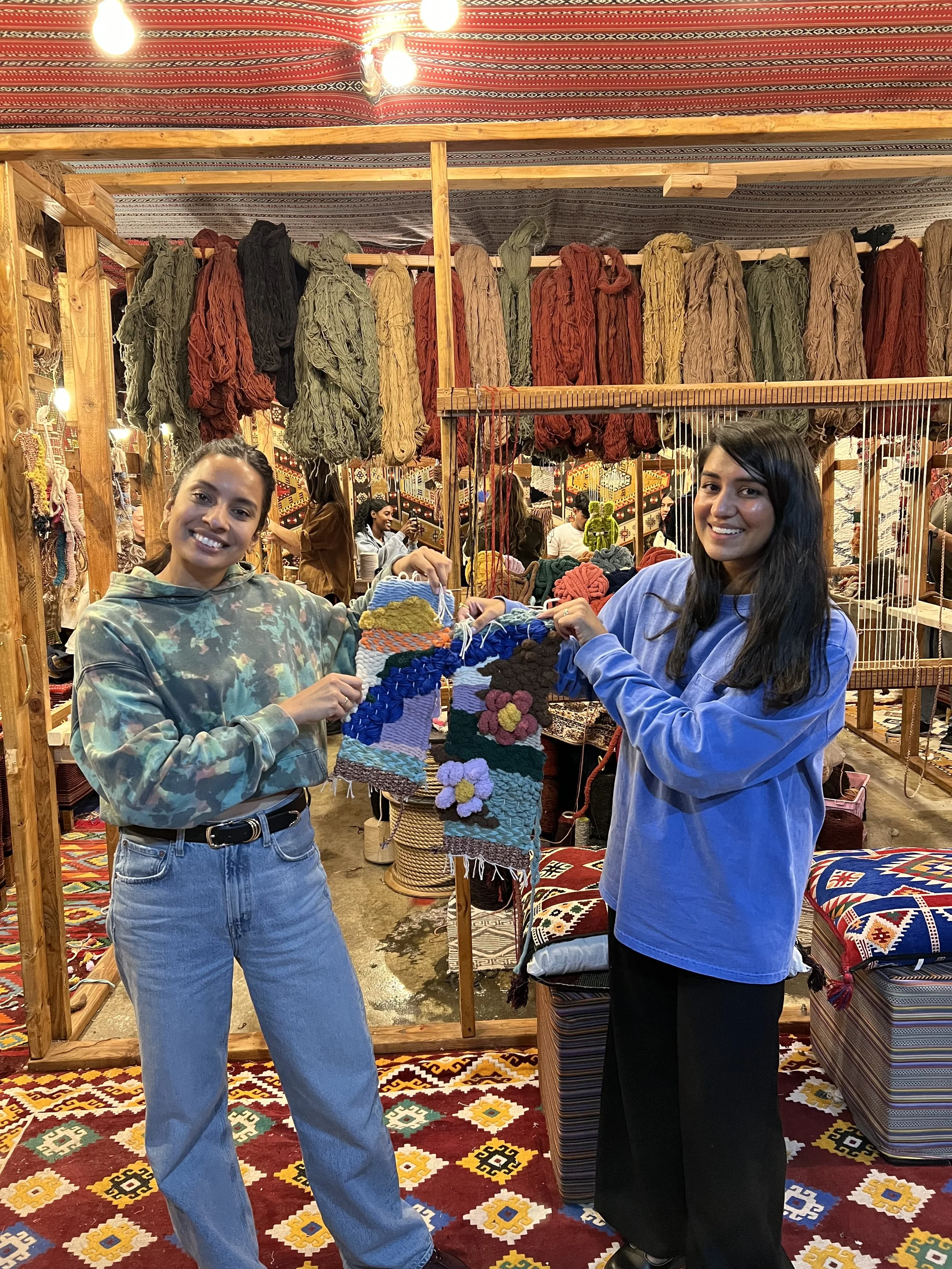 Two women smiling and holding colorful woven wall hangings or rugs inside a craft shop with hanging yarns and textiles in the background.