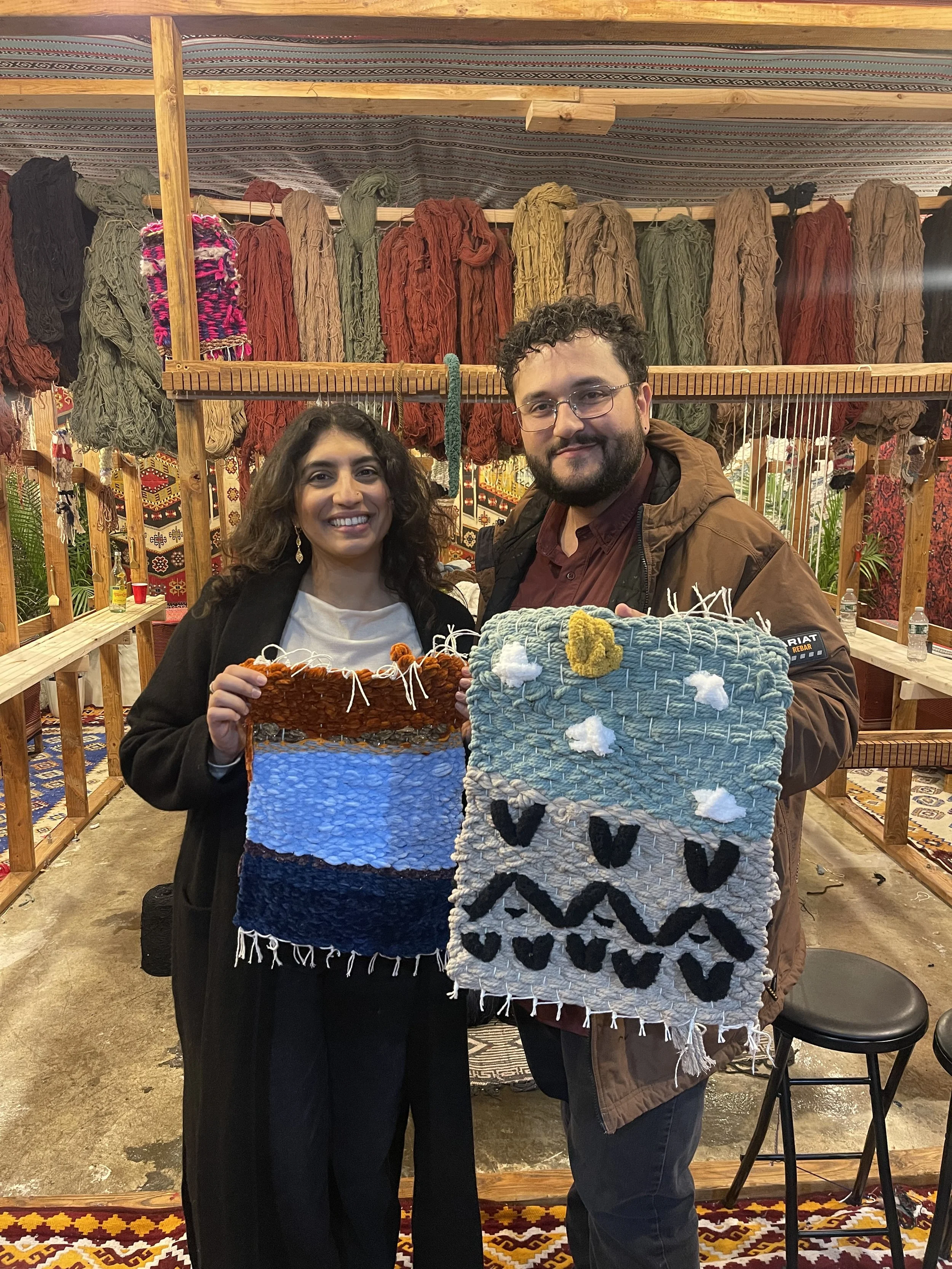Two people smiling and holding handmade woven wall hangings in an indoor craft booth with colorful yarns hanging on the wall behind them.