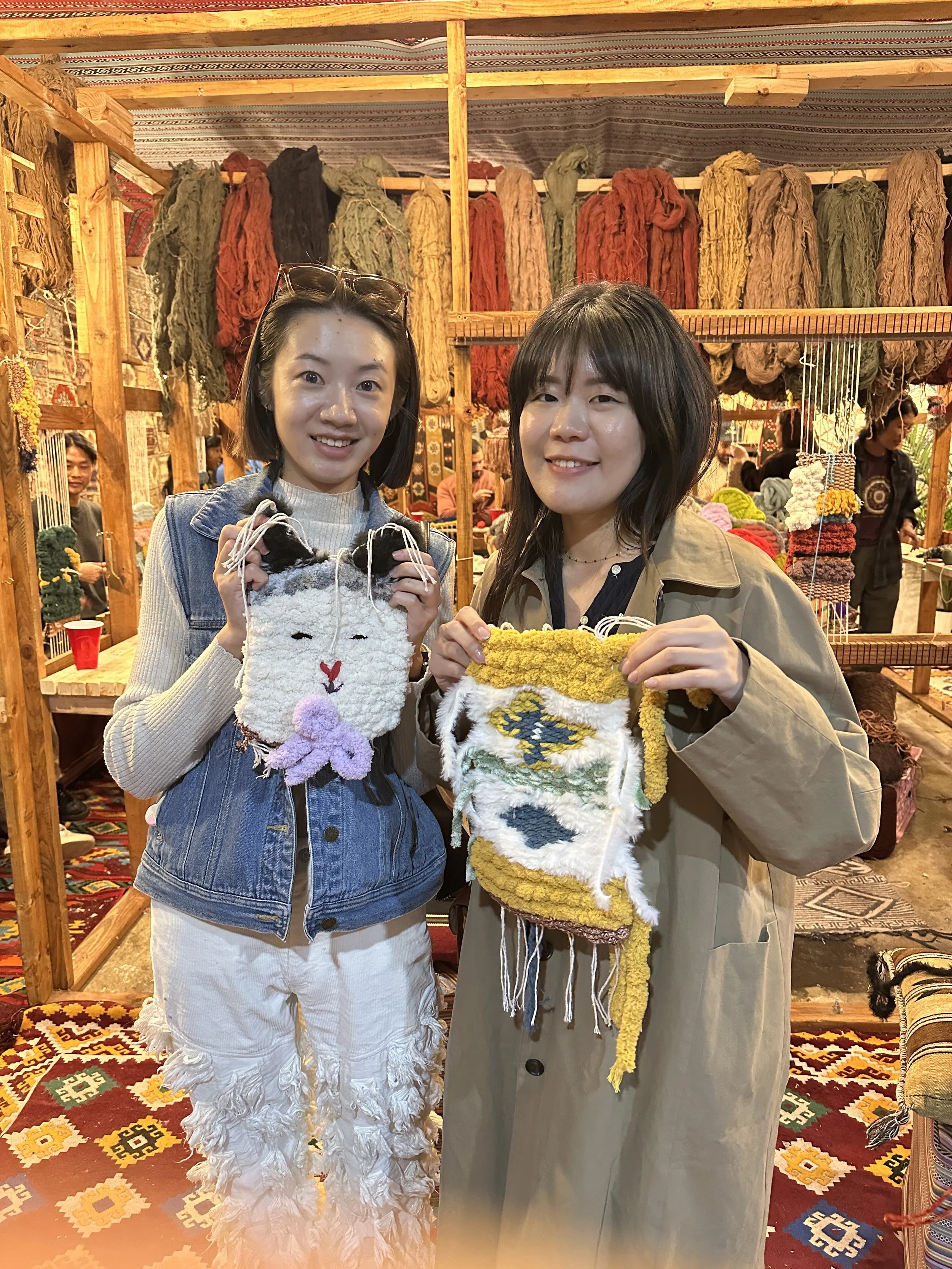 Two women standing inside a craft booth, holding colorful handmade felted bags with animal and abstract designs. The background displays hanging yarn and textile art, with other people also browsing the craft items.