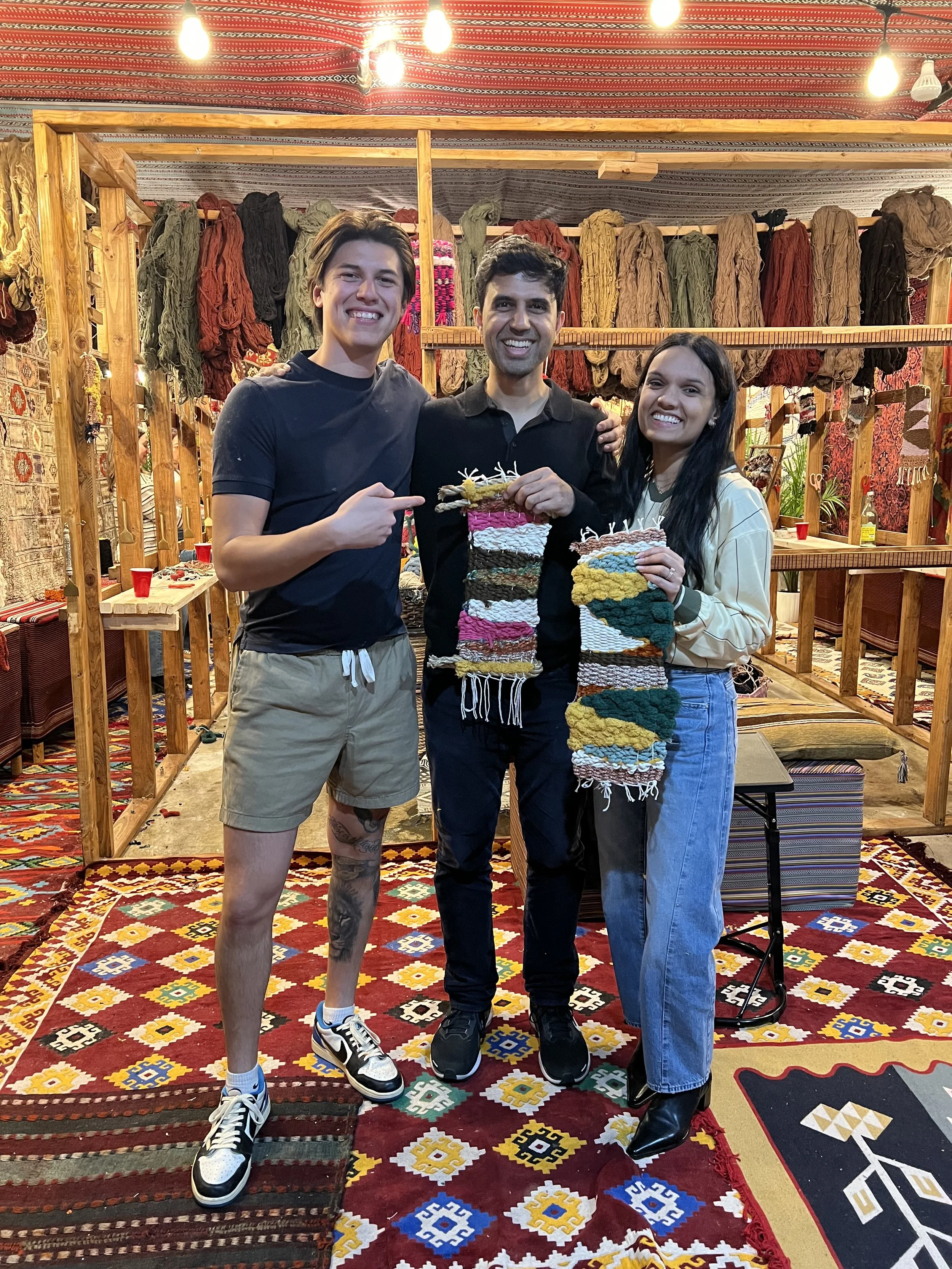 Three people smiling at the camera inside a colorful textile or craft shop, holding small woven rugs, with various yarns and textiles on shelves in the background.