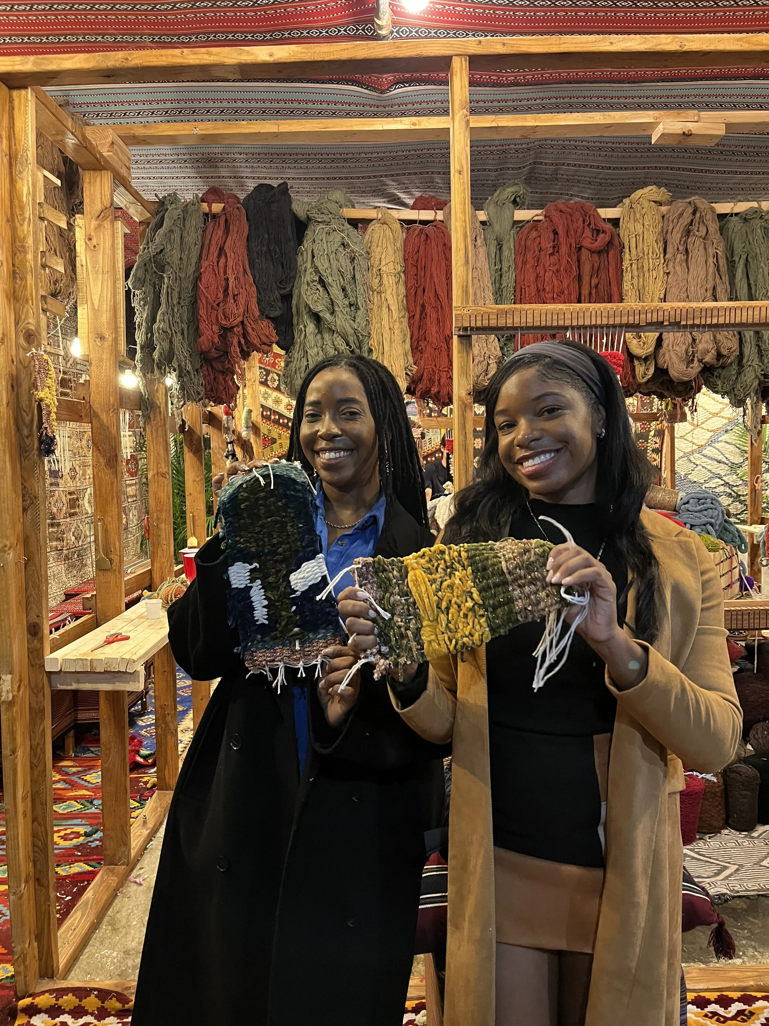 Two women smiling and holding woven textile samples inside a craft store with shelves of yarn and woven items in the background.