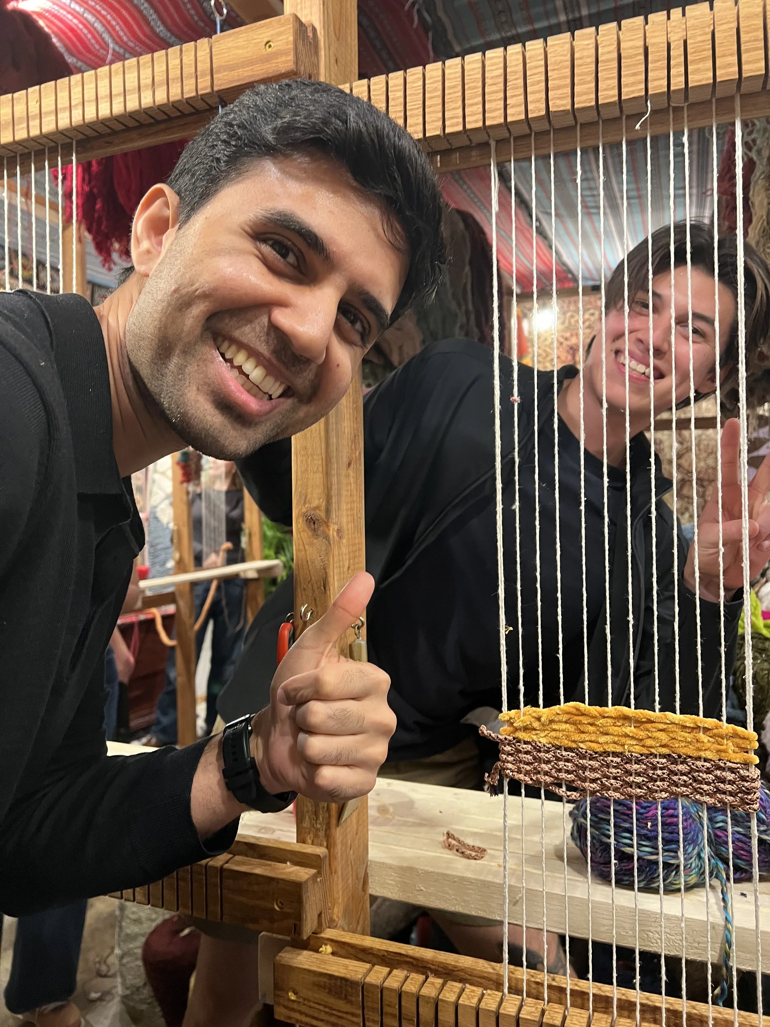 Two smiling men at a yarn weaving booth, one pointing and the other making a peace sign behind a wooden frame with colorful yarn.