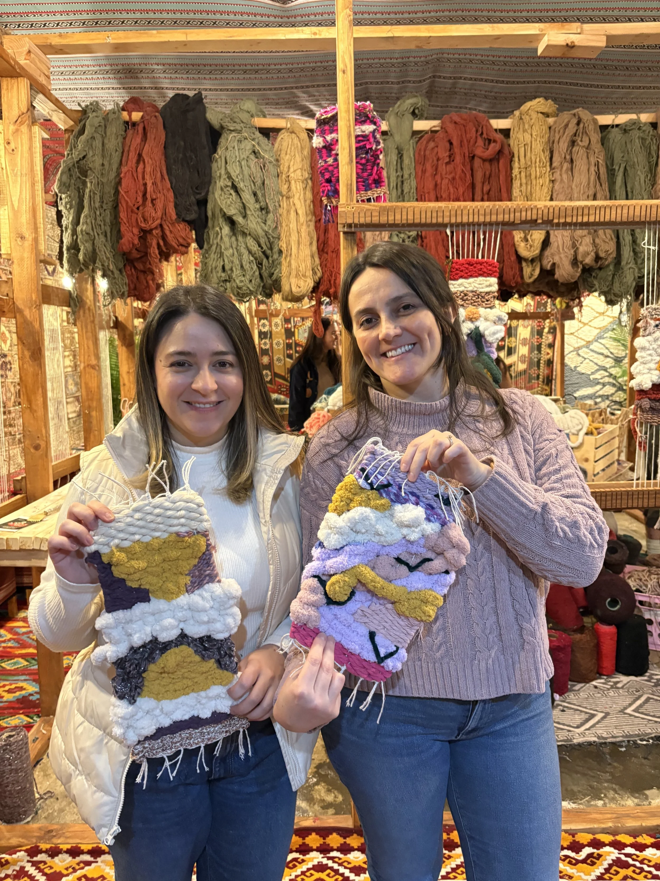 Two smiling women holding colorful woven wall hangings at a market stall with hanging wool yarn in various colors behind them.