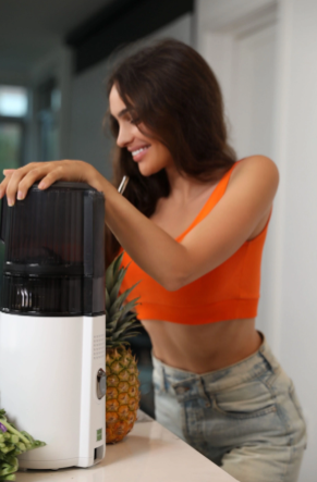 A woman wearing an orange top smiling while using a Hurom juicer on a kitchen counter, with fresh pineapple and vegetables nearby.