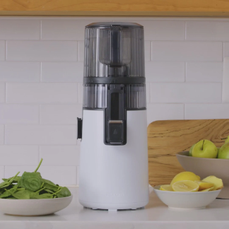 Hurom H70 vertical slow juicer in a modern kitchen setting, placed on a countertop with bowls of fresh spinach, apples, lemon slices, and ginger nearby.